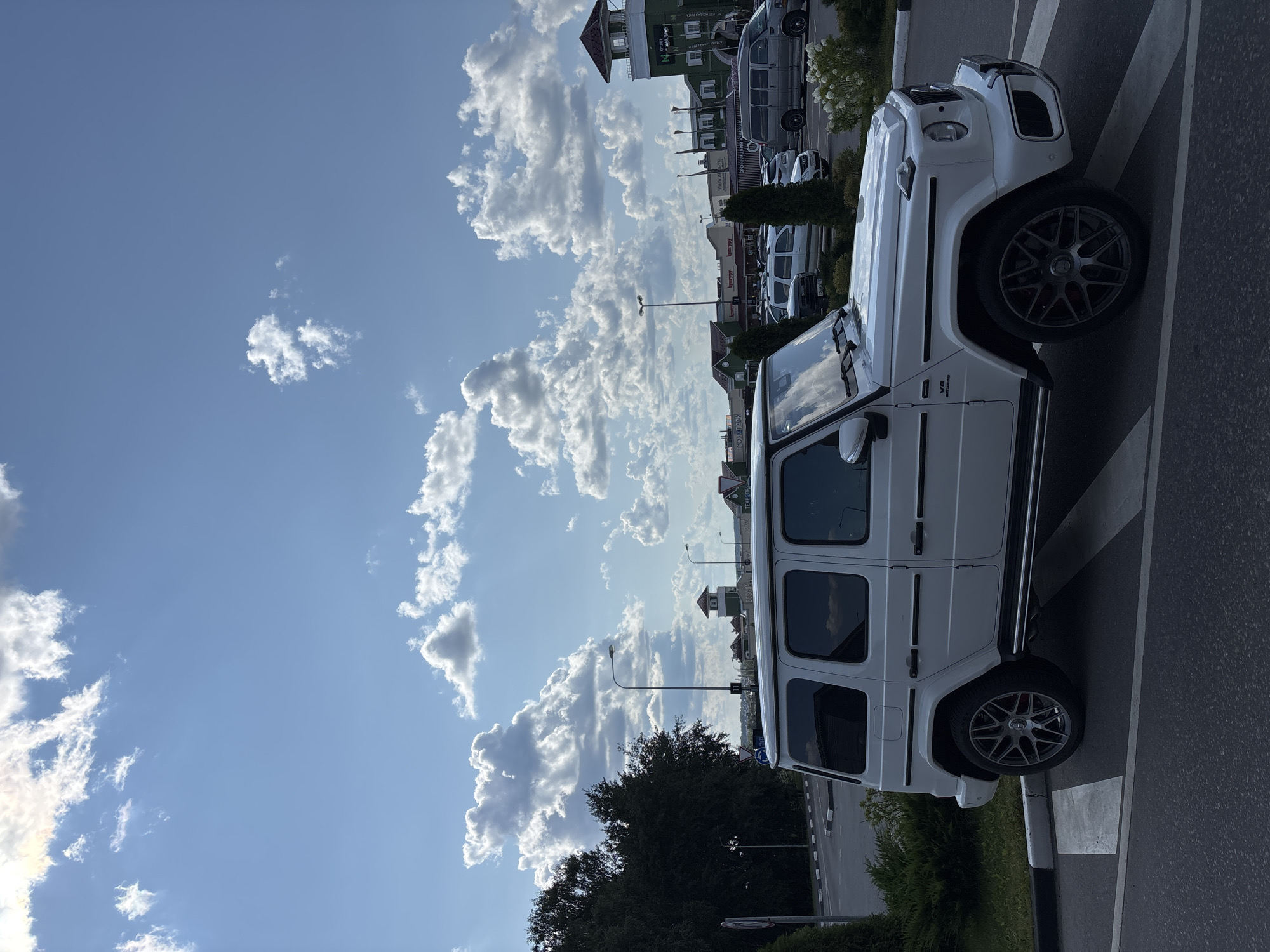 A white Mercedes-Benz G-Wagen is parked in a lot with other vehicles under a partly cloudy sky. The vehicle is a four-door SUV with tinted windows and custom rims. In the background, there are several buildings with shops and a green tower. The scene appears to be in an outdoor shopping area.