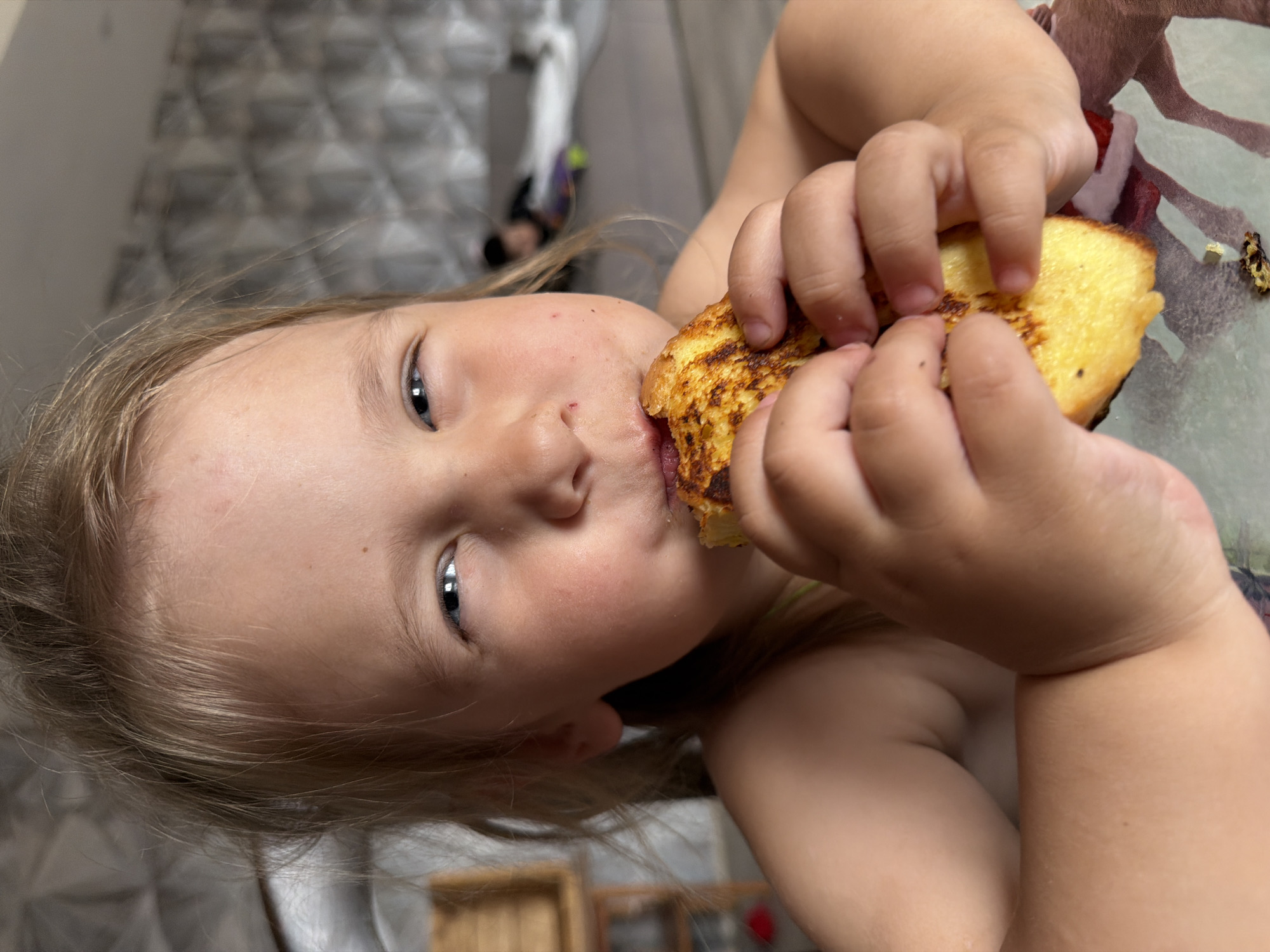 A close-up, high-angle shot captures a young child in an indoor setting in Chaikovsky, Russia, likely during the daytime. The child, with fair skin and light-colored hair, looks towards the camera while holding and eating a piece of food. Their light-colored eyes are prominent, and they appear focused on their meal.
The child is actively biting into a piece of golden-brown food, which resembles fried bread or French toast. Both small hands are wrapped around the food as they bring it to their mouth. Traces of a darker substance, possibly jam or another topping, are visible near the child's mouth and on the food itself, indicating they are in the process of eating.
The background is softly blurred, but hints of a light-colored, geometrically patterned fabric (possibly bedding or a blanket) are visible, suggesting a casual home environment. The lighting appears soft and natural, typical of an indoor daytime scene. The overall impression is one of a child enjoying a snack or meal. There is no visible text in the image.