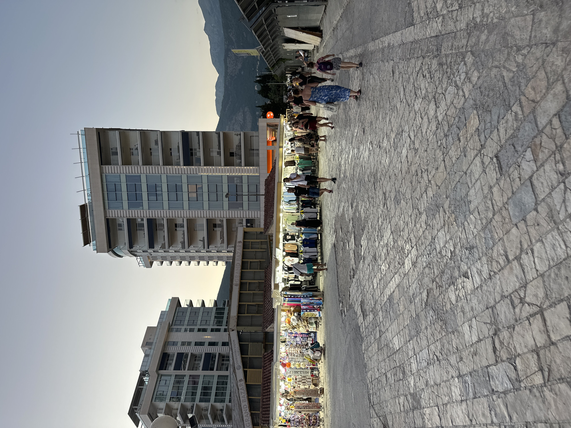 This image depicts a scene in Budva, Montenegro, likely near a shopping area. In the background, two modern, multi-story buildings stand tall against a pale sky, one with a rounded facade and balconies, the other more rectangular. Between the buildings and the foreground, a row of open-air market stalls is illuminated by warm lights, displaying a variety of clothing and accessories, including t-shirts, hats, and souvenirs. Several people are browsing the stalls.
In the foreground, a wide, cobblestone plaza stretches out, with a distinct pattern of irregular stone pavers. Several individuals and small groups of people are walking across the plaza. Some are dressed in casual summer clothing. The lighting suggests it is either early morning or late evening. In the far distance, the silhouette of mountains is visible. To the right of the frame, the market stalls continue, with some featuring protective coverings or awnings. The overall atmosphere is one of a bustling commercial area in a Mediterranean city.