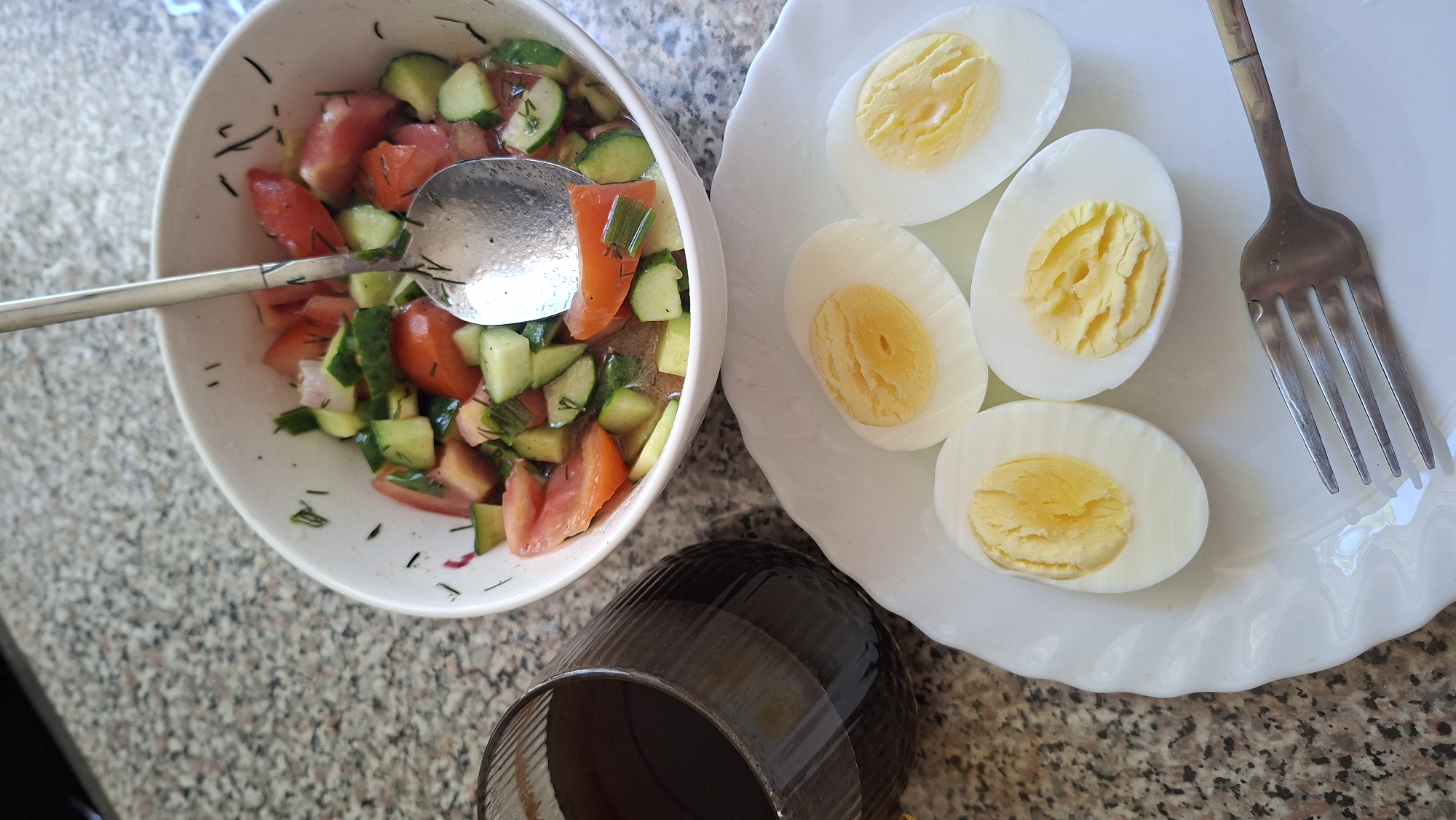 A close-up, top-down view of a meal on a speckled granite countertop. The meal consists of a white bowl filled with chopped tomatoes and cucumbers, seasoned with herbs and a metallic serving spoon resting in the bowl. To the left of the bowl, a dark glass contains a dark beverage. In the foreground, a white plate holds four halves of hard-boiled eggs, with their yellow yolks visible. A silver fork lies on the plate to the right. The scene appears to be a domestic setting, likely a kitchen or dining area. There are no people visible in the image, and no specific landmarks or context are provided to pinpoint the location within Myadzyel, Belarus.