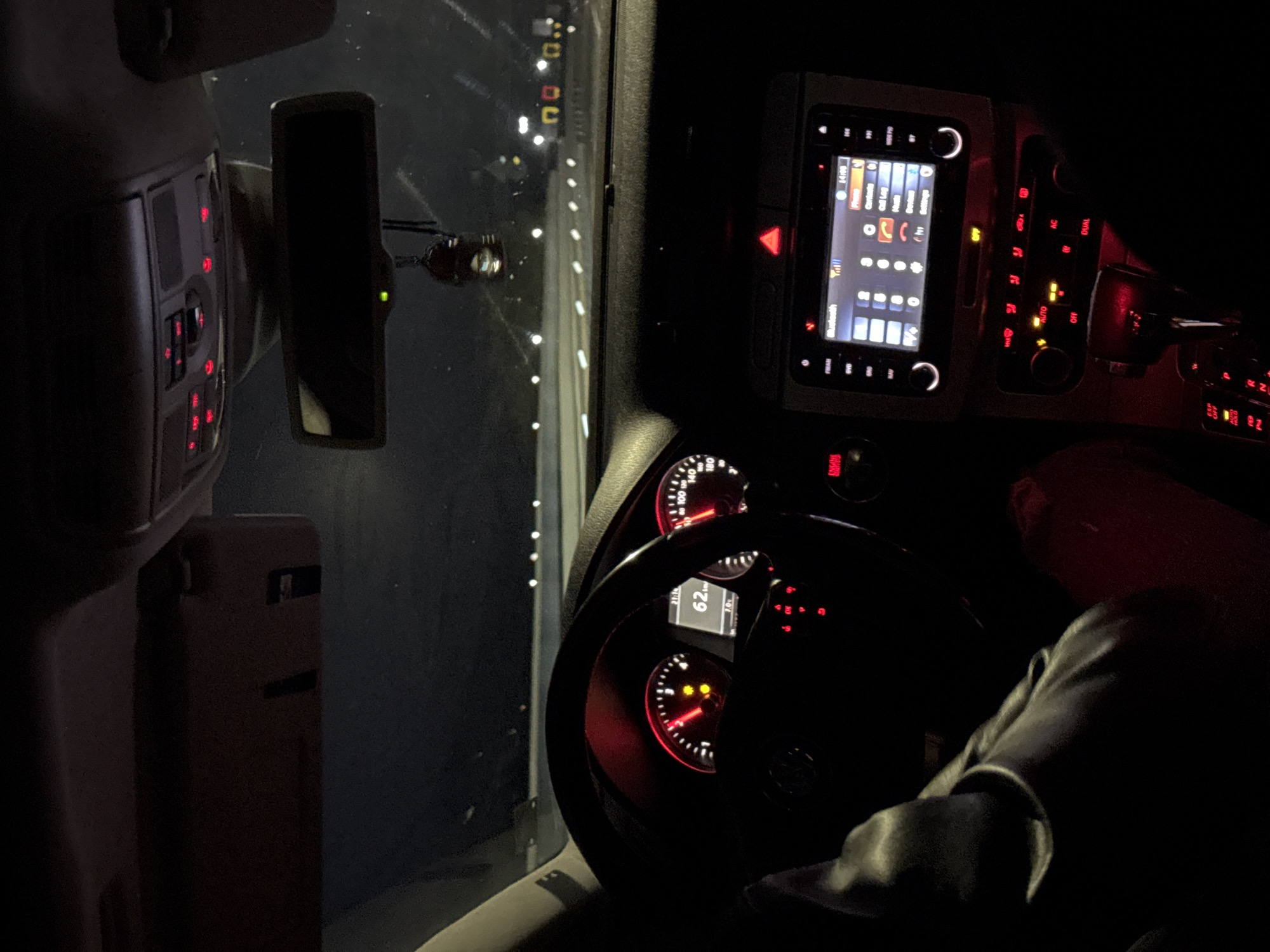 An interior shot of a car at night, taken from the passenger side looking towards the driver's seat and the road ahead. The overall scene is dark, illuminated primarily by the red and white lights of the car's dashboard and infotainment system.
In the foreground, part of a person's leg or arm, clad in dark clothing, is visible to the right. The steering wheel occupies the center-left, with the instrument cluster visible through it. The speedometer shows "62", likely indicating the vehicle's speed in kilometers per hour, alongside other markings like "140" and "180". A digital display within the cluster shows "21:19" (time) and "7.0". Red warning lights and indicators are active on the dashboard.
To the right of the steering wheel, the central console features an infotainment screen displaying a phone interface with options like "Phone", "Contacts", "Call Log", "Bluetooth", and "Settings". A time of "14:08" is shown on this screen, and icons for signal strength and battery are visible. Below the screen, the climate control panel shows illuminated buttons with text such as "AUTO", "AC", "OFF", and "DUAL". A hazard light button (red triangle) is also illuminated. Further right, a key can be seen in the ignition.
An overhead console with red illuminated buttons is visible in the upper left, next to the rearview mirror. Outside the front windshield, a dark environment with distant, scattered white lights suggests the car is either on a road at night or parked in a dimly lit area, possibly in Barakovo, Bulgaria. The rearview mirror shows a faint green light.