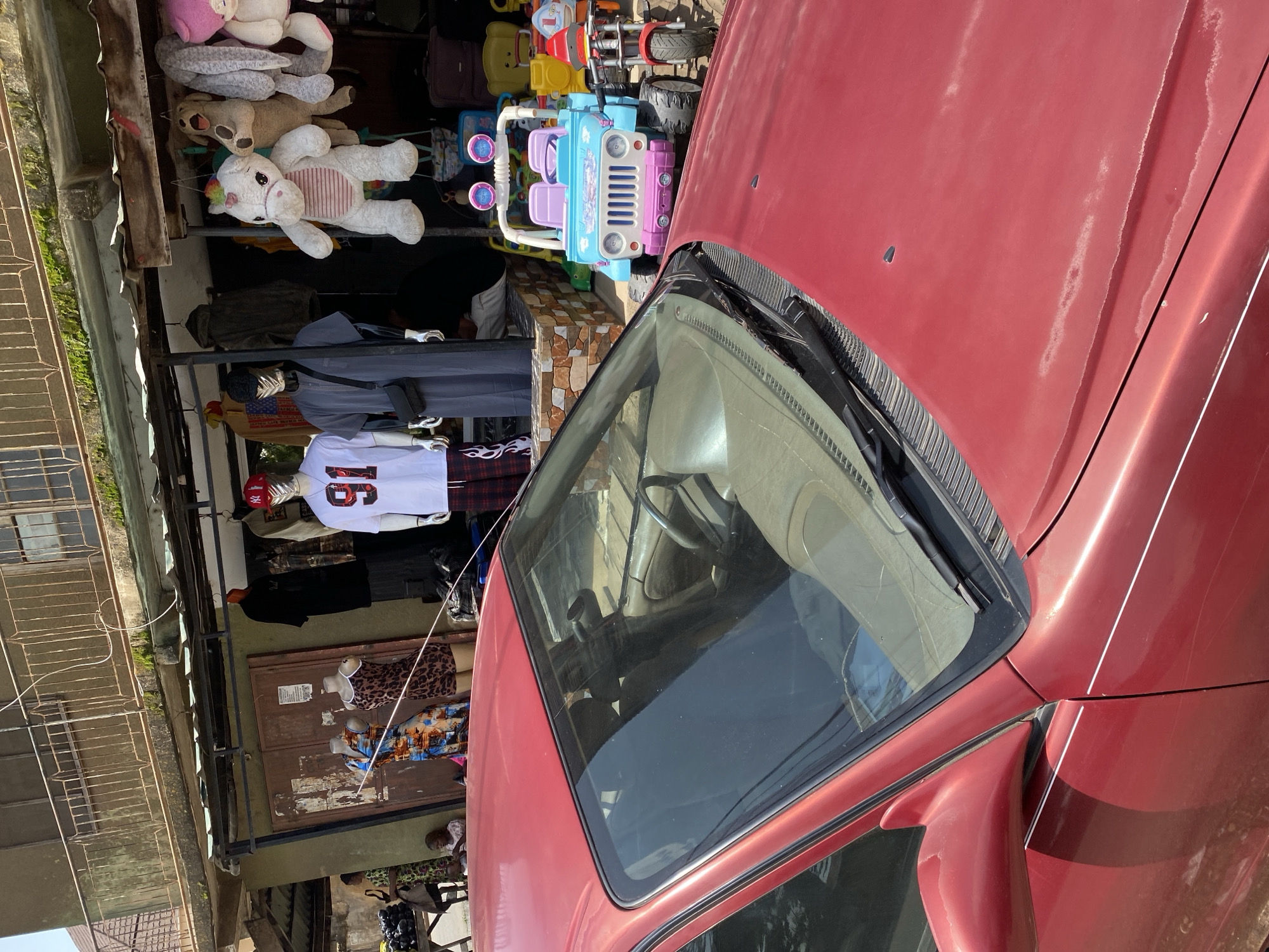 The image depicts an outdoor market scene in Jos, Nigeria, with a red car prominently in the foreground. The car's hood and windshield are visible, suggesting it is parked in front of a stall. Behind the car, a market stall is set up with various items for sale. These include clothing displayed on mannequins, with one mannequin wearing a white t-shirt with the number "91" and a red baseball cap, and another mannequin dressed in a gray robe with a crossbody bag. Other clothing items are hung on racks. Further back, several stuffed animals, including a white unicorn and a brown dog, are displayed. To the right, children's ride-on toys are visible, specifically a blue and pink jeep and a red toy motorcycle. A woman in a patterned dress is seen in the background, near a pile of what appears to be produce. The market appears to be situated in a urban area, with a building structure visible behind the stalls. The overall impression is a busy marketplace.