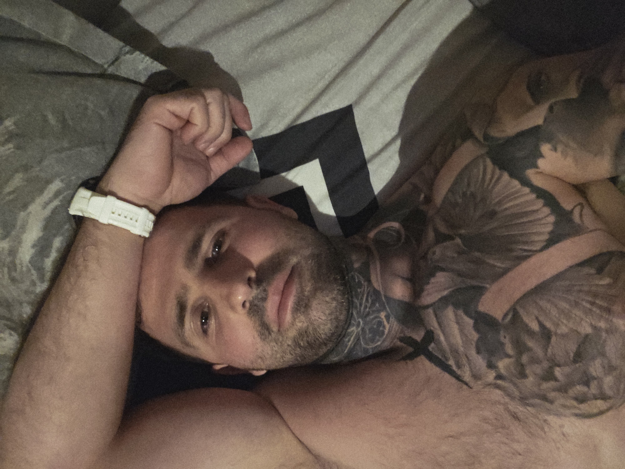 A man with tattoos on his chest and neck is lying on a bed in Stara Zagora, Bulgaria. He is wearing a white watch on his left wrist and his right arm is bent and resting on his head. The bed is covered with a grey sheet with a black and white geometric pattern. The lighting in the room is dim.