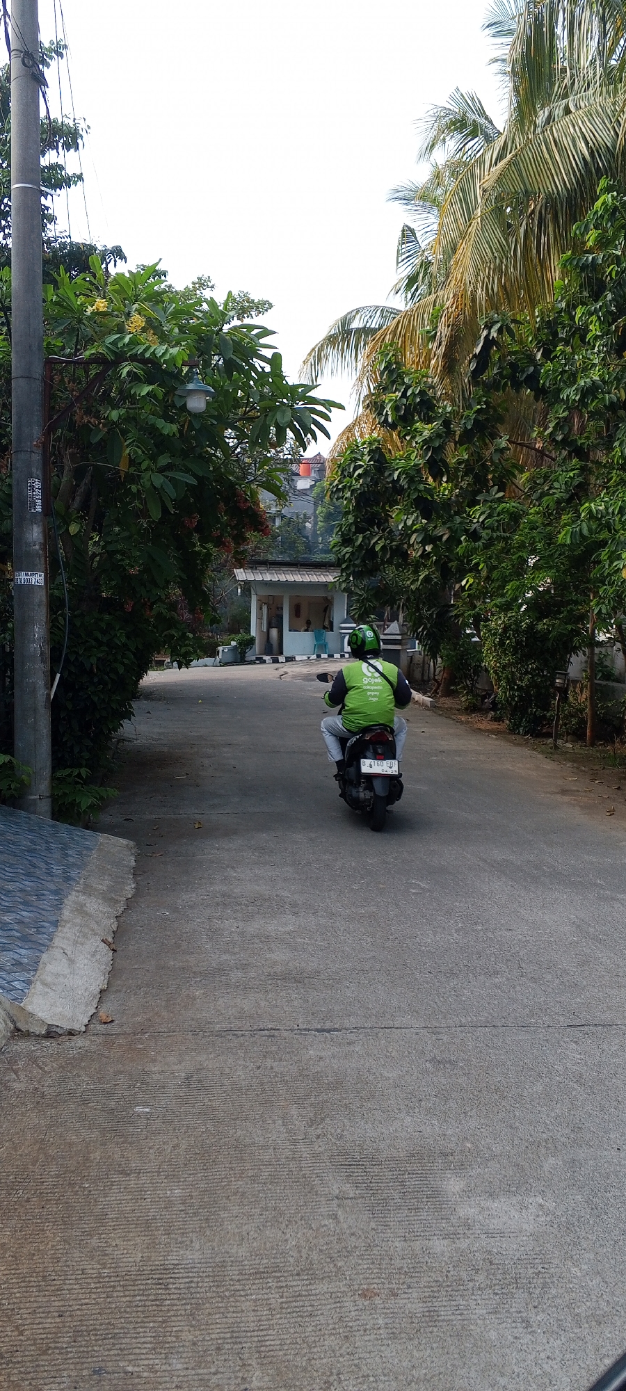 In Depok, Indonesia, a person wearing a green Gojek uniform rides a motorcycle down a paved road lined with trees and foliage. The motorcycle has a license plate that reads "B 4160 EBF". In the background, a small building with a white roof and blue trim is visible, with two chairs outside. A utility pole stands to the left of the road, and the sky is overcast.