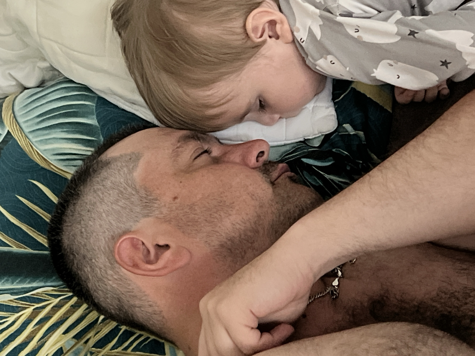 A close-up shot features an adult male lying down, possibly on a bed, alongside a young child. The child, clad in a gray pajama top patterned with white sheep and stars, rests their head on a white pillow. The man's head is positioned adjacent to the child's, his eyes closed. He has short, graying hair and facial hair. The bedspread is decorated with a botanical design of green and gold palm fronds. A silver chain necklace is visible on the man's chest. The location of the image is unknown and cannot be determined to be in the city of Noviy, Russia.
