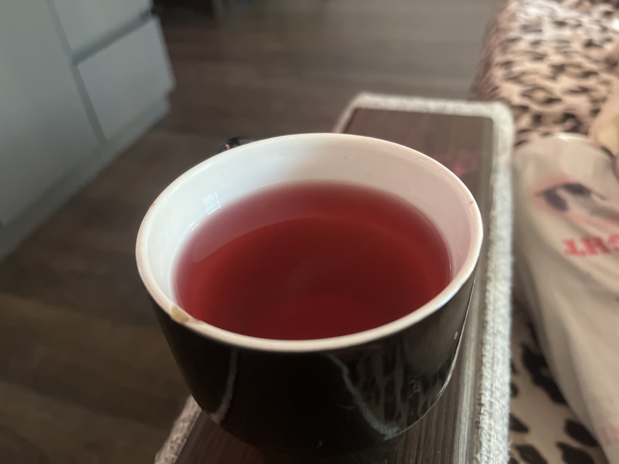 A close-up shot depicts a black and white marble-patterned mug filled with a red liquid, likely tea or juice. The mug is positioned on a dark surface with a textured, possibly crocheted, edge. To the right of the mug is a partially visible object, potentially a bag, with a pattern or text visible. The background includes a piece of furniture with drawers and a blurred pattern that appears to be animal print. The setting seems to be indoors, possibly a living room or office. There are no people visible. The location information cannot be determined from the given image.