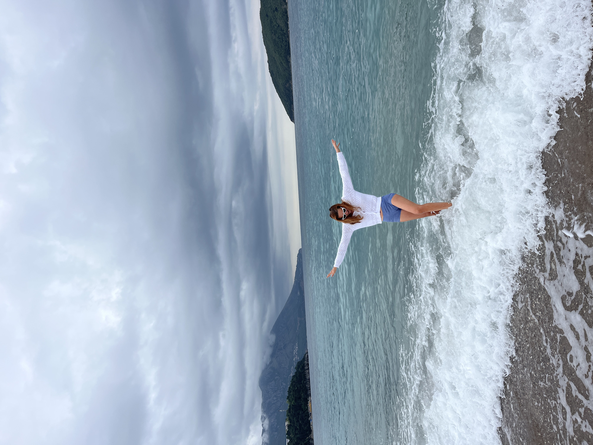 An outdoor photograph, originally oriented vertically but depicting a horizontal scene, shows a woman standing at the edge of a body of water with dramatic mountains in the background under an overcast sky. The woman, who has long hair and is wearing a white long-sleeved top, blue shorts, and white sunglasses, stands with her arms outstretched horizontally, suggesting a joyful or free pose. Her feet and lower legs are immersed in the foamy white waves that break on the shore.
The water to her left is a deep blue-green, showing subtle ripples across its surface. The shoreline, visible beneath the breaking waves, appears to be composed of pebbles or dark sand. In the distant background, rugged mountains rise directly from the coast, their peaks partially shrouded by low-hanging, grey clouds. The sky is entirely overcast, indicating a cloudy day, yet the scene is well-lit. The combination of mountains, sea, and beach suggests a dramatic coastal setting, consistent with locations like Budva, Montenegro. There is no visible text in the image.