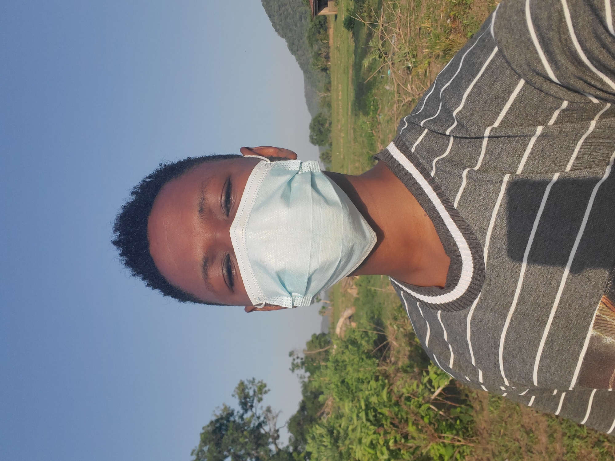 A close-up, portrait-oriented photograph shows a person wearing a light blue surgical mask and a gray-and-white striped shirt. The person is facing to the left, with their face partially obscured by the mask. Only one eye is clearly visible. The background includes foliage and trees, with a hill or mountain range visible in the distance. The sky is light blue, suggesting daytime. The location appears to be an outdoor area in Mararaba, Nigeria.