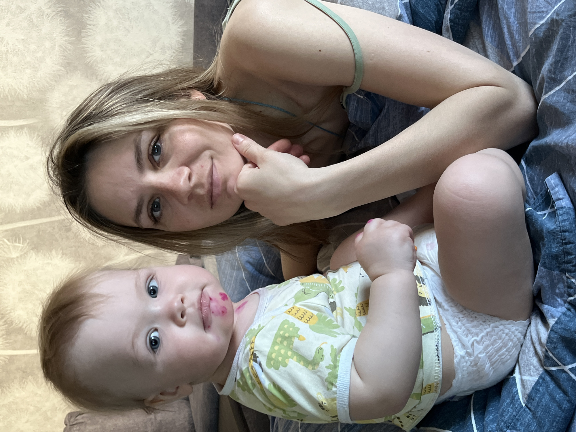 A woman and a baby are lying on a bed. The woman has blond hair and blue eyes, and is wearing a light green tank top. She is touching her chin with her hand. The baby has reddish blond hair and blue eyes, is wearing a patterned yellow and green onesie, and a diaper. There are red marks around the baby’s mouth. The bed has blue and gray striped bedding.
While the image shows a woman and baby, there is no indication of the city Chaikovsky, Russia, or location information within the image.