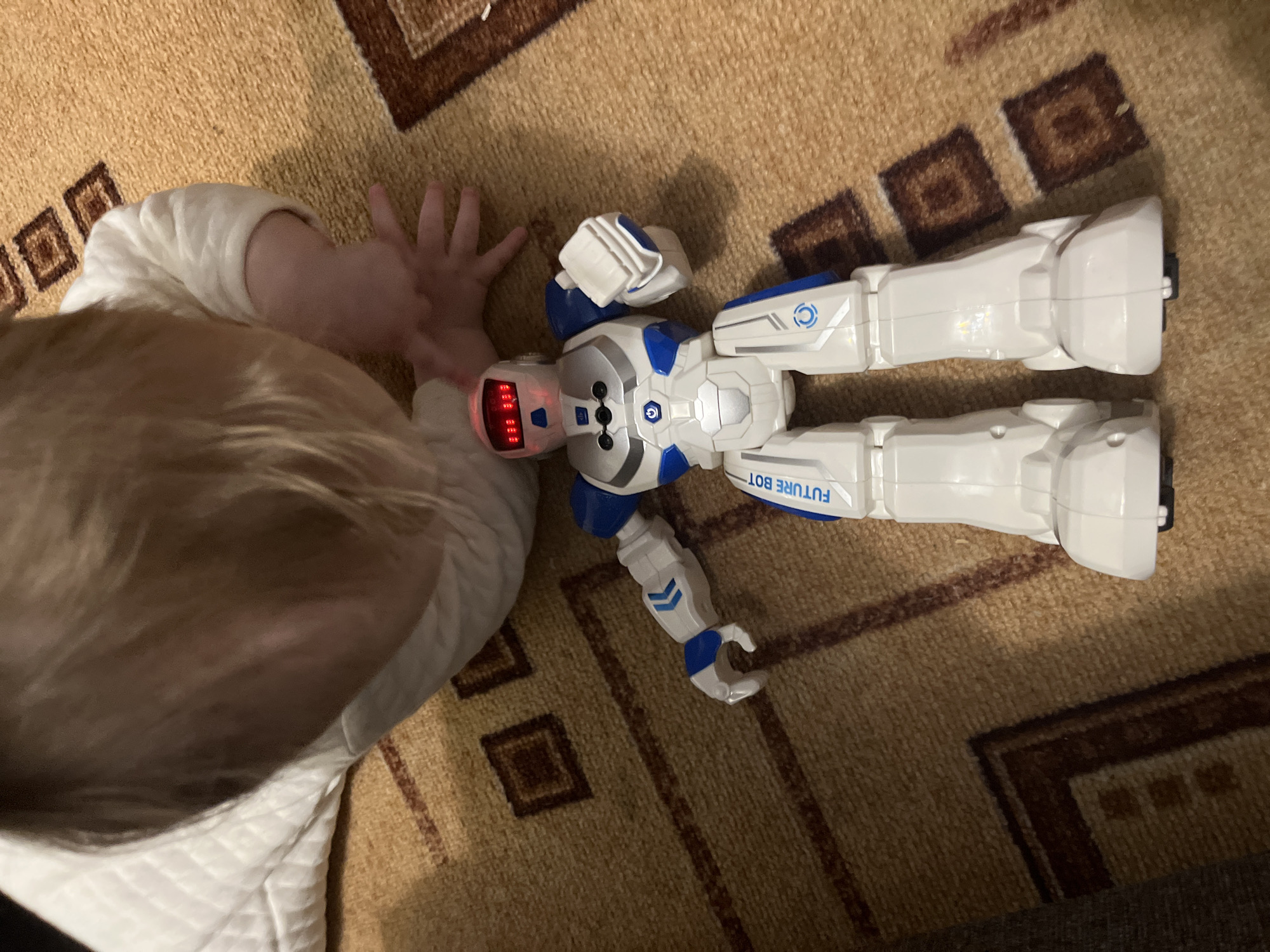 An overhead view depicts an indoor scene in Chaikovsky, Russia, featuring a child interacting with a toy robot on a patterned carpet. A child, identifiable by light-colored hair and an arm in a white quilted garment, is positioned on the floor, with their right hand extended towards the robot. The robot, primarily white with blue and silver accents, is labeled "FUTURE BOT" on its left leg and lies face-up on the light brown carpet. Its face displays red illuminated eyes and a red-lit mouth-like grid. The carpet features a repeating pattern of dark brown geometric shapes, including squares and borders.