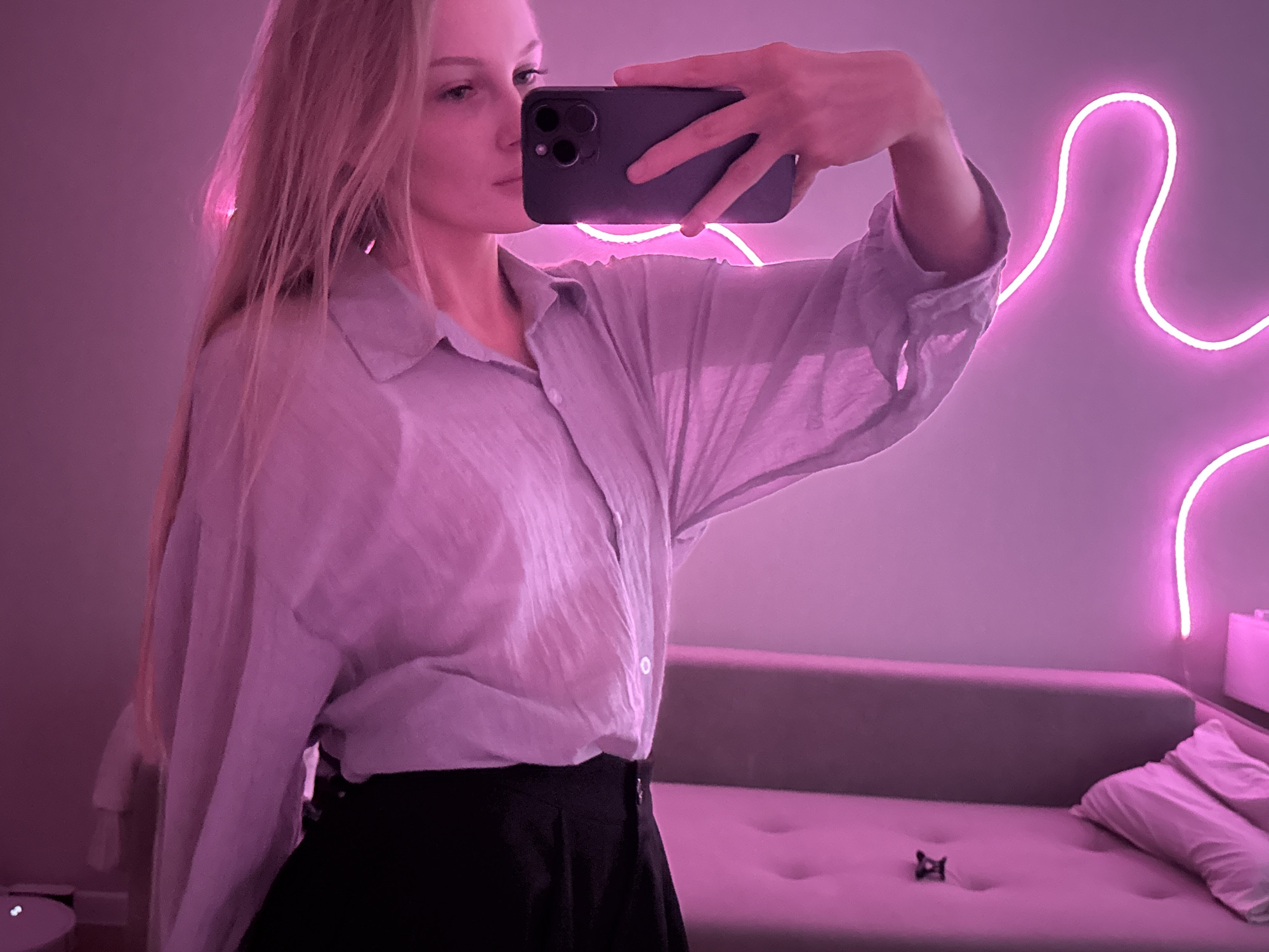A young woman with blonde hair is taking a selfie in a room with pink neon lighting. She is wearing a light-colored, slightly sheer button-down shirt and black bottoms. Her phone is visible in her right hand as she holds it up to take the photo. In the background, a sofa is visible, as is a pink neon light installation on the wall. There are no discernible landmarks or objects in the image that would indicate a specific location within the city of Perm, Russia. The scene is indoors and appears to be a personal living space.