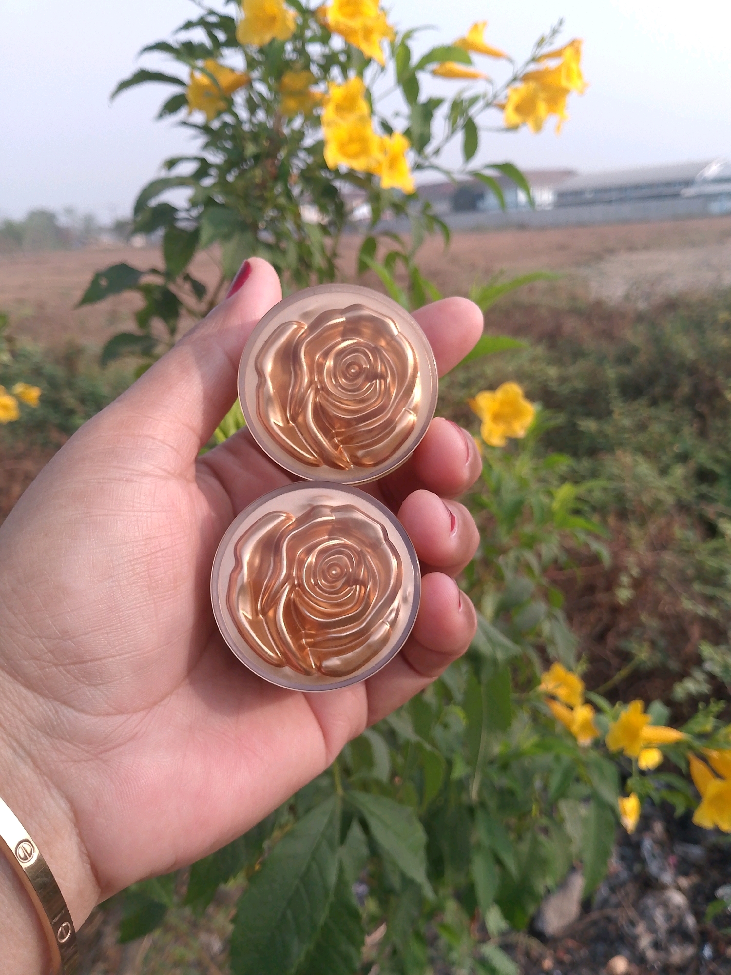 A person's hand, adorned with a gold bracelet, holds two round, rose-embossed compacts against a backdrop of blurred yellow flowers and greenery. The compacts appear to be makeup products. The scene is outdoors, possibly in a rural or suburban area, given the natural elements and a distant building. The image was captured in Cisait, Indonesia.