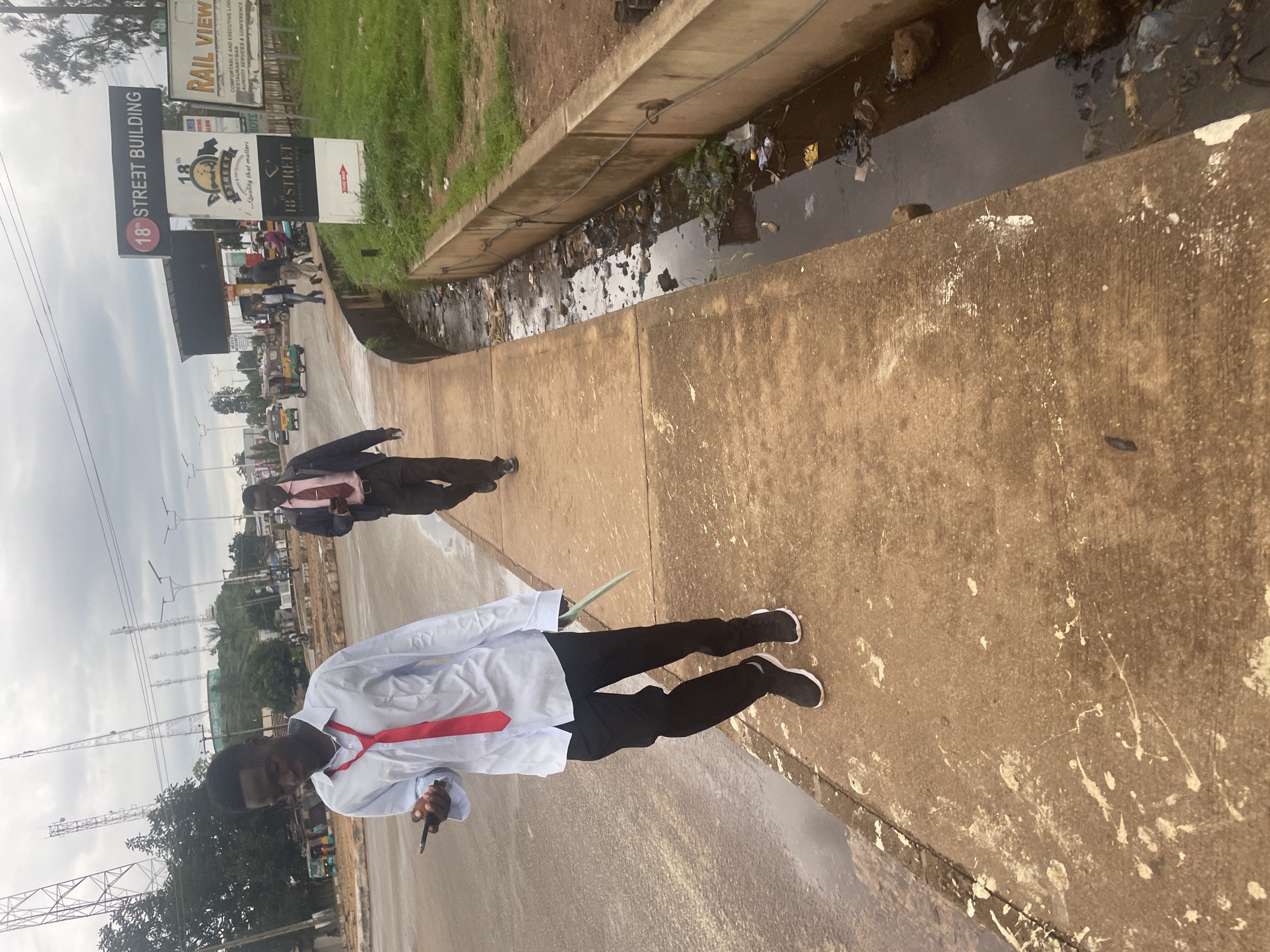 In the city of Jos, Nigeria, two individuals are observed walking along a concrete sidewalk adjacent to a drainage ditch filled with refuse. The person in the foreground, a young man, wears a light blue long-sleeved shirt, a red tie, black trousers, and black and white athletic shoes. He is looking down at his mobile phone, which he holds in his left hand, and carries a light green flat object in his right hand. Behind him, another man in a pink shirt, dark jacket, and dark trousers is also walking, looking at his phone. In the background, a busy street is visible with several vehicles, including a three-wheeled auto-rickshaw. Various signs are present, including "18th STREET BUILDING" and "RAIL VIEW". The environment appears overcast with visible utility poles and structures in the distance, suggesting an urban setting. The drainage ditch is heavily polluted with plastic waste and other debris.