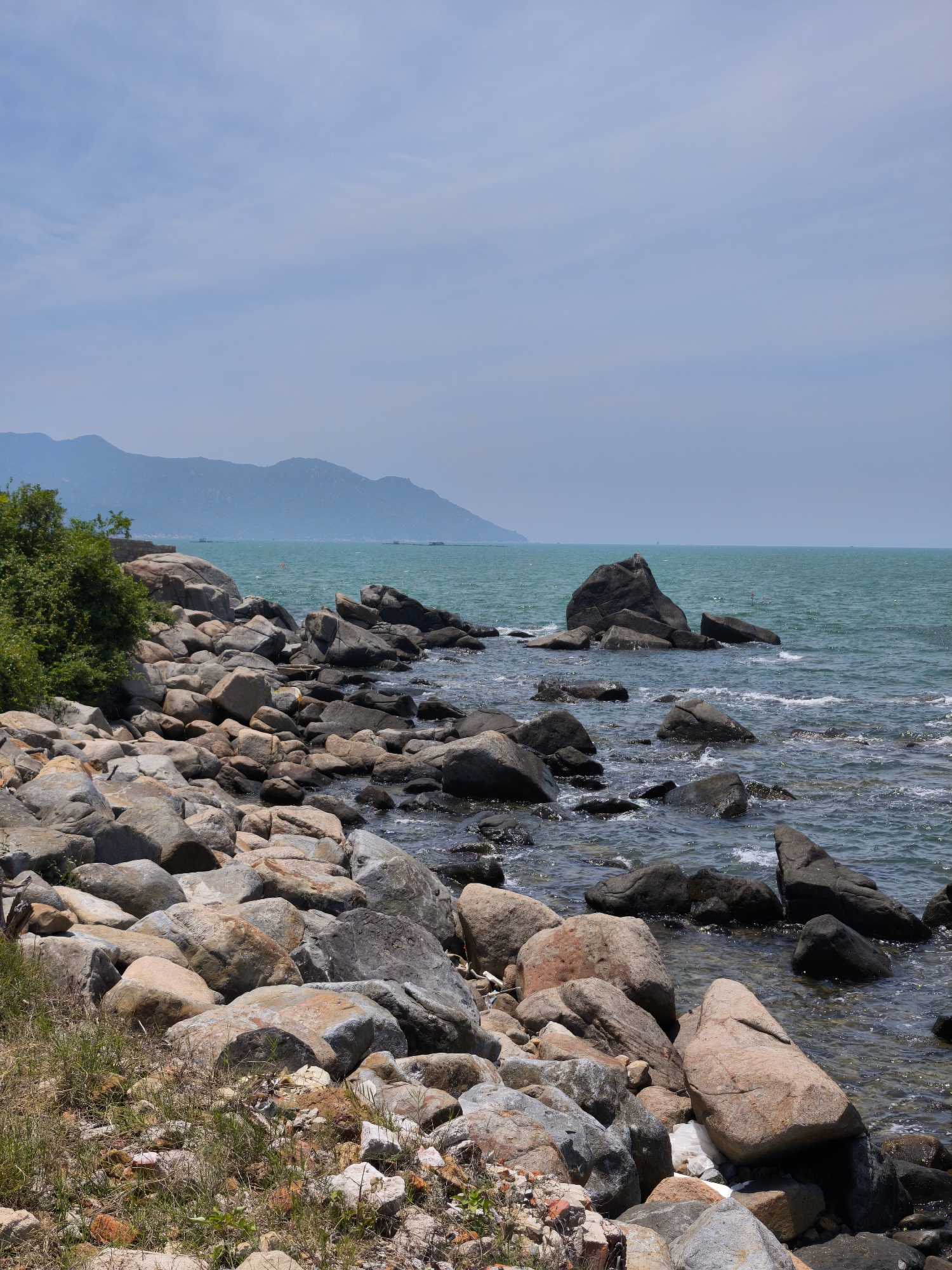 The image depicts a rocky shoreline in Vĩnh Lương, Vietnam. The foreground is dominated by a dense collection of large, weathered rocks of varying shapes and colors, ranging from grey and brown to pinkish hues. Patches of dry grass and sparse green vegetation grow among the stones.
In the middle ground, the rocks extend into the turquoise-colored ocean, with waves gently lapping against them. A cluster of dark, prominent boulders juts out of the water, creating a more dramatic seascape. Further out, the ocean stretches to the horizon under a pale blue sky with wispy clouds.
In the background, a range of hazy, blue-green mountains forms the distant coastline. A few indistinct structures or boats can be seen on the water near the mountains. The overall impression is one of a natural, slightly rugged coastal environment. There are no people or animals visible in the image.