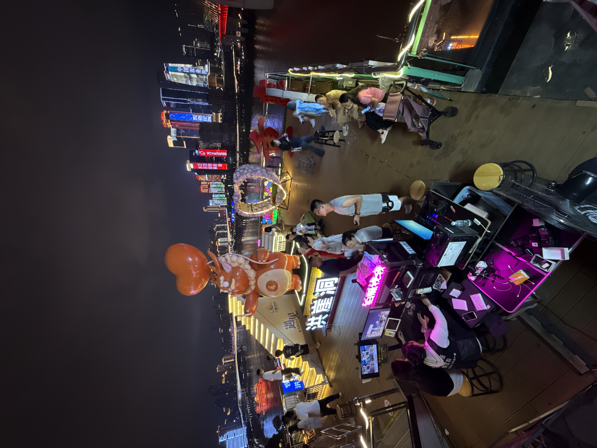 The image depicts a nighttime scene on a boat or floating platform docked in the Yuzhong District of Chongqing, China. The backdrop features a panoramic view of illuminated skyscrapers lining the riverfront, reflecting in the dark water. In the foreground, a group of people are gathered on a wooden deck. To the left, two individuals are seated at a desk with multiple computer monitors displaying graphics and text, suggesting a gaming or streaming setup, bathed in purple and pink neon light.
Further back, a large, orange, cartoonish mascot with heart-shaped balloons stands near a brightly lit staircase leading up to a higher level of the platform. Several people are seen walking up and down these stairs, while others are congregating or sitting on the deck. To the right, a heart-shaped decorative structure adorned with lights and flowers is visible, possibly a photo opportunity. Various tables, chairs, and a stroller are also present on the deck, indicating a public gathering space. The overall atmosphere is lively and illuminated by a combination of city lights, neon signs, and decorative lighting on the platform itself.