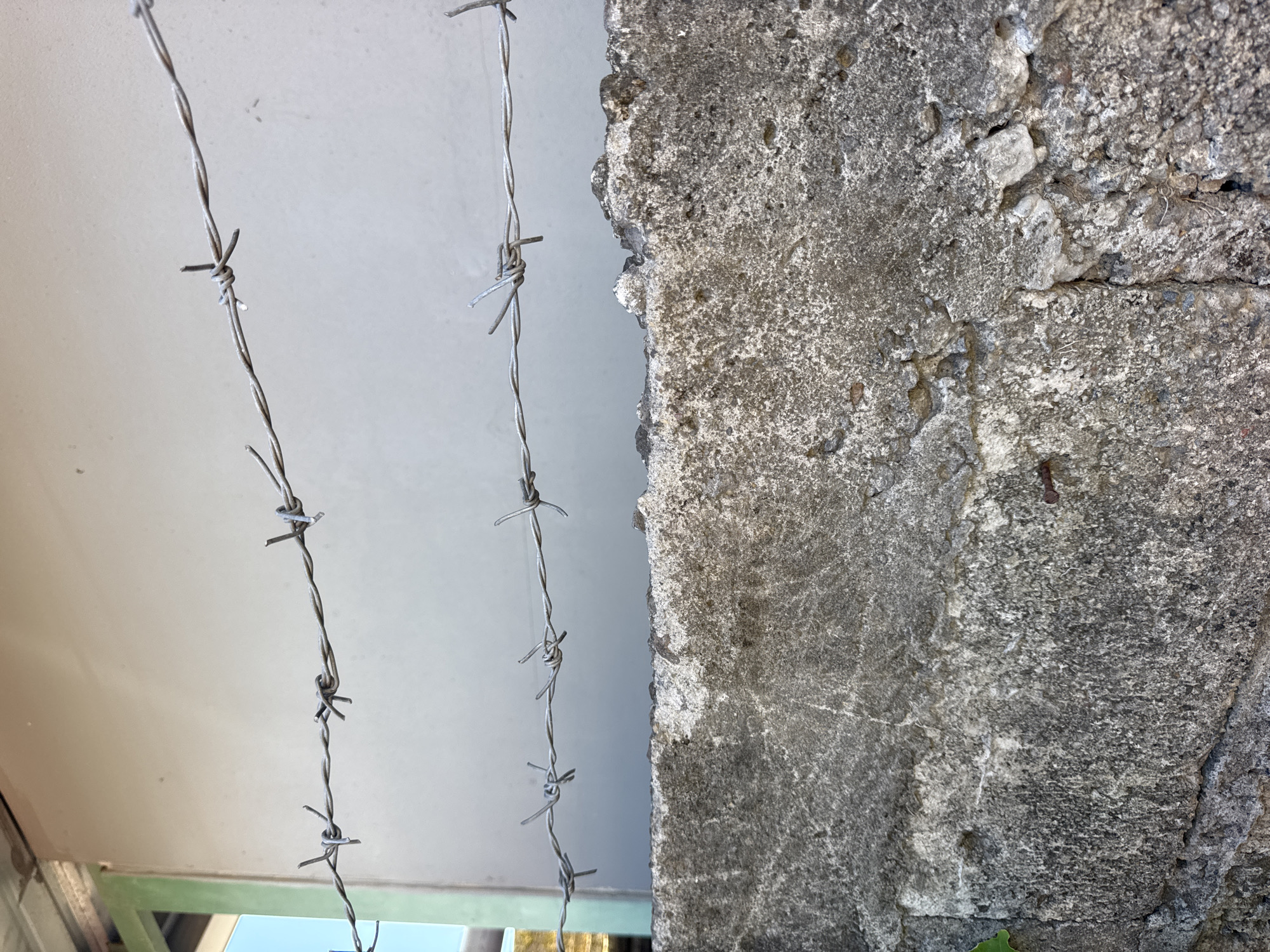 A close-up shot shows two strands of barbed wire hanging vertically near a rough, grey concrete structure. The wire is silvery, and the barbs are sharp and regularly spaced. The concrete surface is weathered and uneven, with visible cracks and textures. A glimpse of a light-colored ceiling and a window frame are visible in the lower-left corner, indicating the scene might be indoors or under a shelter. The location, Kuta Selatan, Indonesia, is not evident from the visual elements alone. No people, subjects, actions, or interactions are visible in the image.