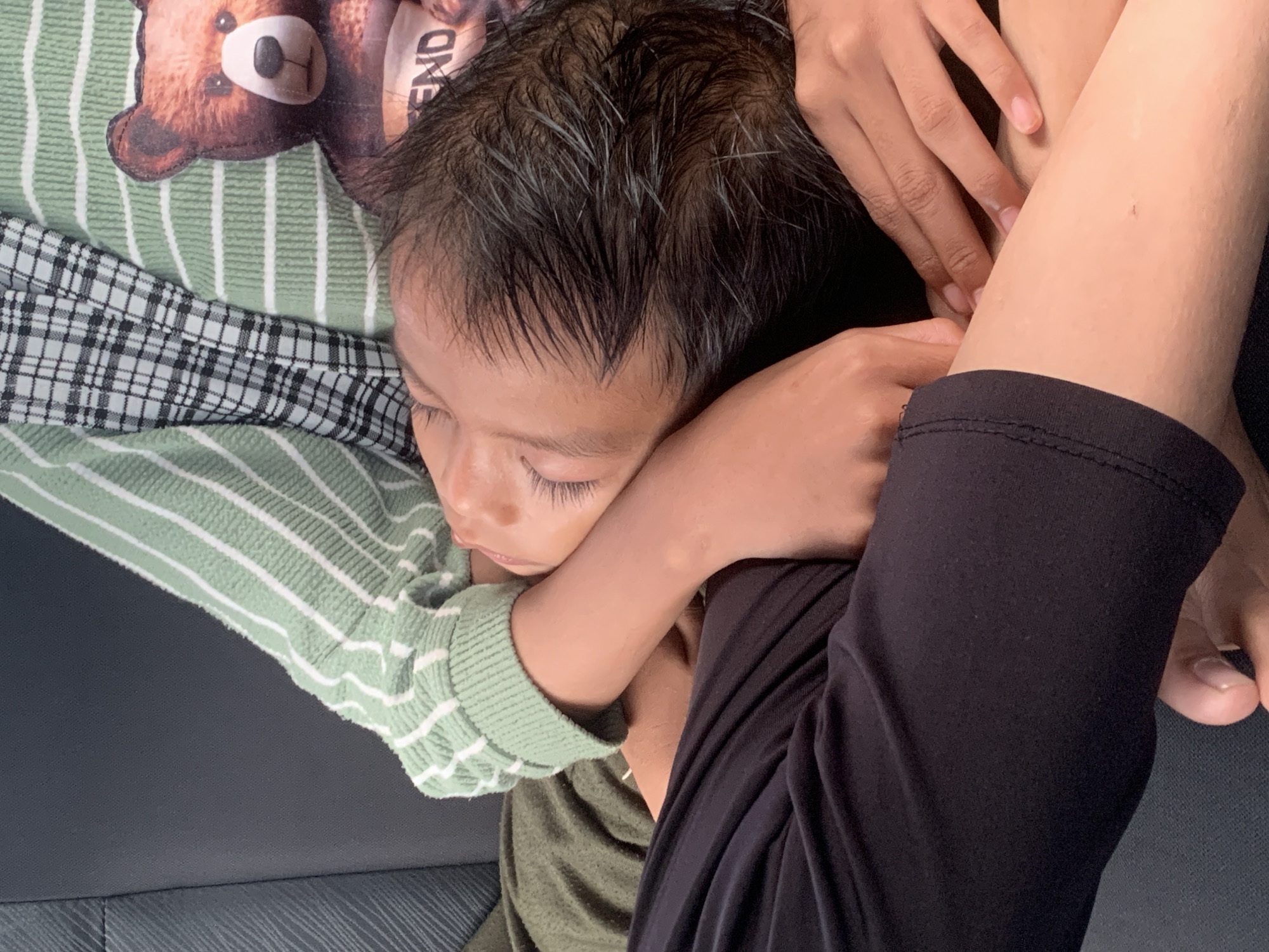 A child is sleeping, nestled on or next to someone. The child has dark hair and closed eyes. They are wearing a green and white striped sweater. The person next to the child is wearing black pants or leggings and has a hand resting near the child's head. A portion of a plush toy with a bear on it is visible. The background appears to be the interior of a car, on a grey seat. There is no information to suggest the location in Samarinda, Indonesia.