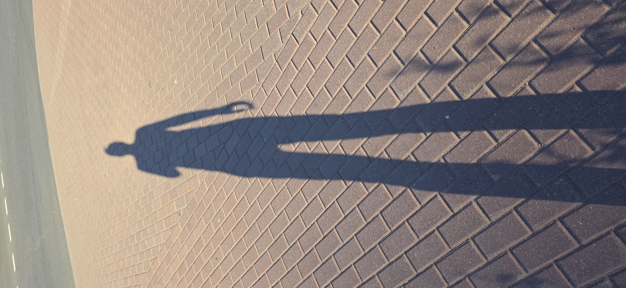 A long shadow of a person is cast on a brick-paved surface adjacent to a road. The person is facing away from the camera and their shadow extends diagonally across the pavement. The shadow's head is visible, along with their torso, arms, and legs. One arm appears to be bent at the elbow, and the person's hand is not clearly defined. The shadow is elongated, suggesting the sun is at a low angle. The road in the background has white dashed lines marking lanes. The overall scene is an outdoor urban environment.