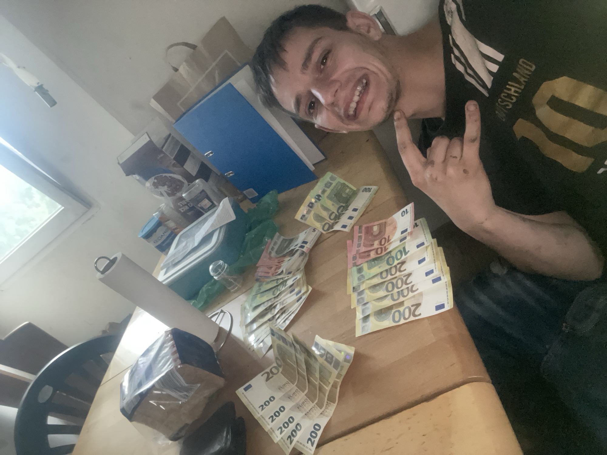 A young man is sitting at a table with money spread out before him. He is wearing a black t-shirt with "Deutschland" and the number "10" printed on it in gold. He is smiling and making a hand gesture. The table is cluttered with various items, including a package of bread, a roll of paper towels, plastic containers, and folders. The room appears to be a domestic setting, possibly a kitchen or dining room. The lighting suggests it is daytime.