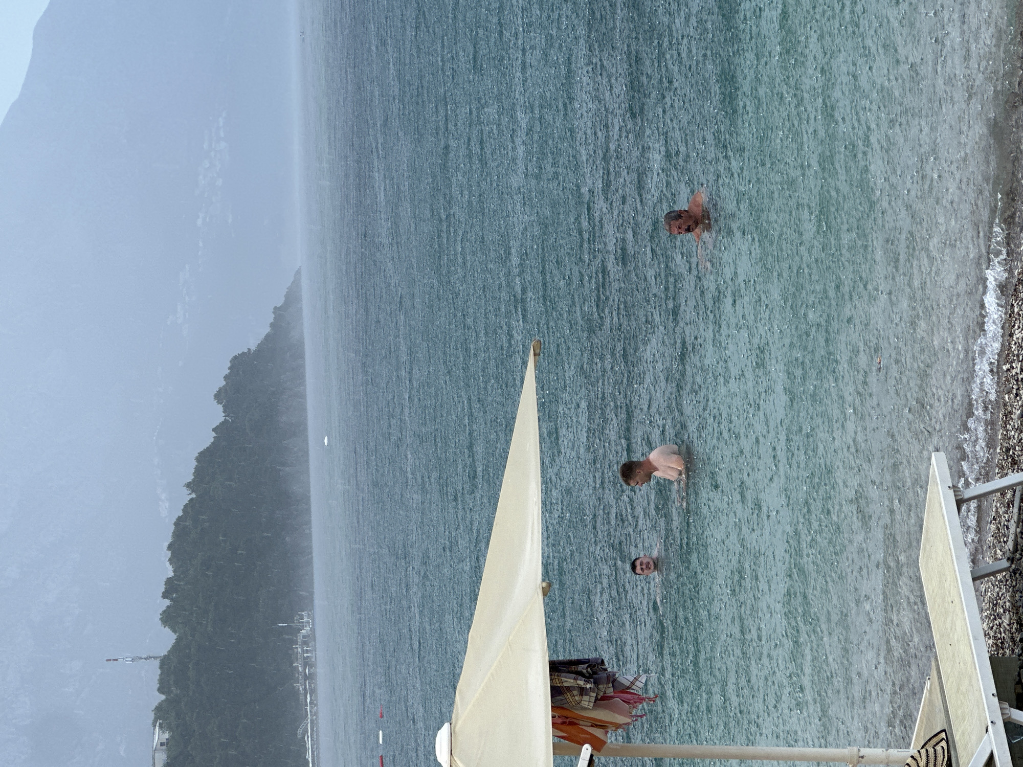 In this media file, the scene depicts a beach in Budva, Montenegro, likely during a downpour, as evidenced by the visible rain streaks across the water. Three individuals are in the sea, with one person closer to the shore and two further out. The water is a murky teal color, indicating turbidity due to the rain. In the foreground, a beach umbrella, partially visible, casts a shadow. To the left, a lush green, tree-covered hill rises steeply from the water's edge, disappearing into a misty, hazy sky. The overall atmosphere suggests a challenging weather condition, yet the presence of people in the water hints at a resilient or perhaps forced continuation of activities. The visibility is reduced, especially towards the distant hills, which are obscured by the weather. The time of day is not clearly discernible due to the overcast conditions.