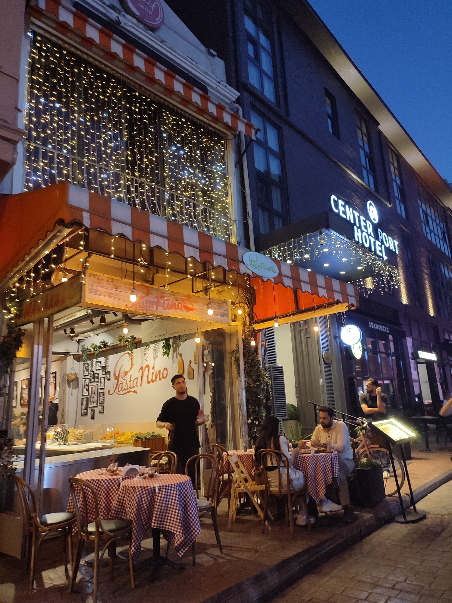 This is a medium shot taken at dusk of an outdoor seating area for a restaurant named "Pasta Nino" on a street in Beyoğlu, Turkey. The restaurant has a striped awning and is decorated with strings of warm white fairy lights. Two tables are set with red and white checkered tablecloths. A male server wearing a black t-shirt and apron is standing inside the restaurant's open-fronted area. A couple is seated at a table to the right, eating dinner. In the background, the "CENTER PORT HOTEL" sign is visible on an adjacent building. The street is paved with cobblestones.