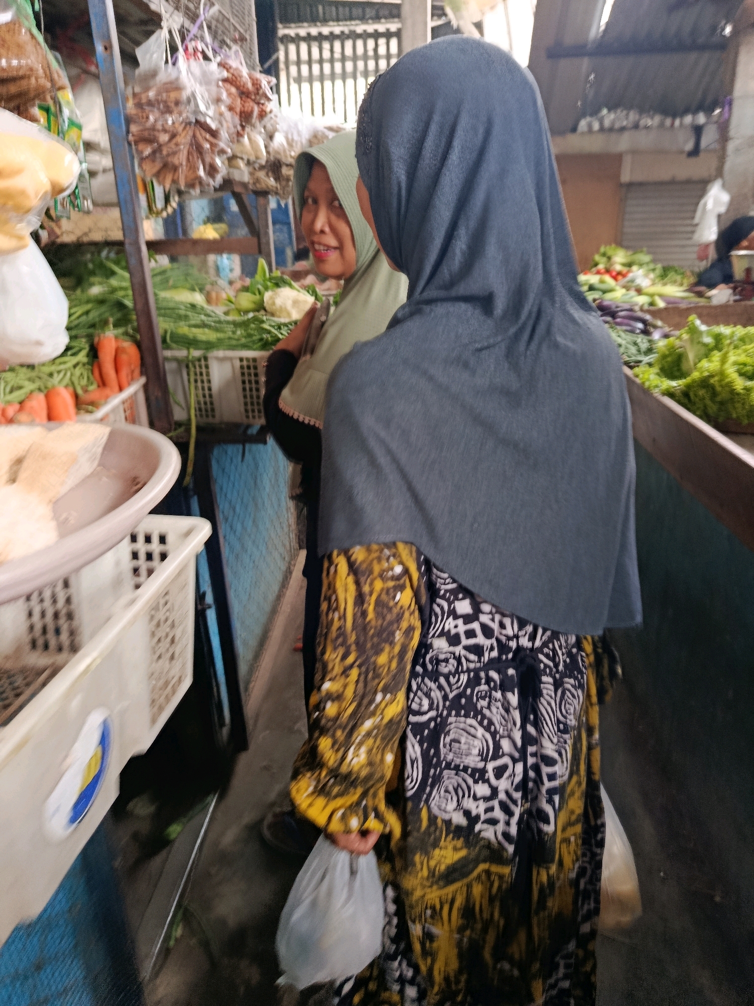 In an outdoor market stall in Bululawang, Indonesia, two women are visible. The woman in the foreground, wearing a dark grey hijab and a patterned dress with yellow and black designs, is seen from the back, holding a plastic bag. To her left, another woman in a light green hijab is partially visible, looking towards the camera. The stall displays a variety of fresh produce, including carrots and green vegetables, arranged in baskets and crates. The background shows market structures and other goods.
