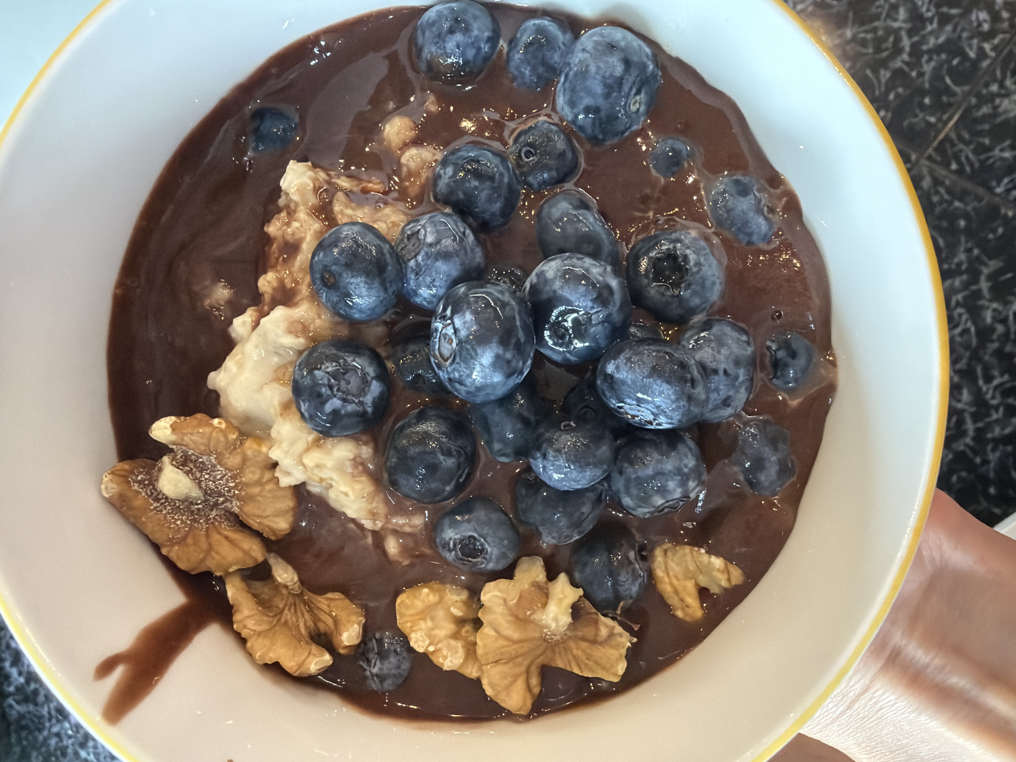 This is a close-up, overhead shot of a bowl of oatmeal topped with blueberries and walnuts, covered in a chocolate sauce. The bowl is white with a yellow rim and sits on a dark, speckled surface. The oatmeal appears to be textured, with visible grains. The blueberries are a deep blue-purple, some glossy and some with a powdery bloom. Several walnut halves are scattered amongst the blueberries and oatmeal, adding a contrasting brown hue and crunchy texture. The chocolate sauce is a rich, dark brown, with visible sheen and ripples, suggesting a creamy consistency. The lighting appears to be natural and bright, casting subtle shadows and highlighting the textures of the food. There is a hand visible on the right edge of the frame, holding the bowl, indicating someone is about to eat or has just prepared the meal. The overall impression is one of a delicious and healthy breakfast or snack. There is no visible text in the image.