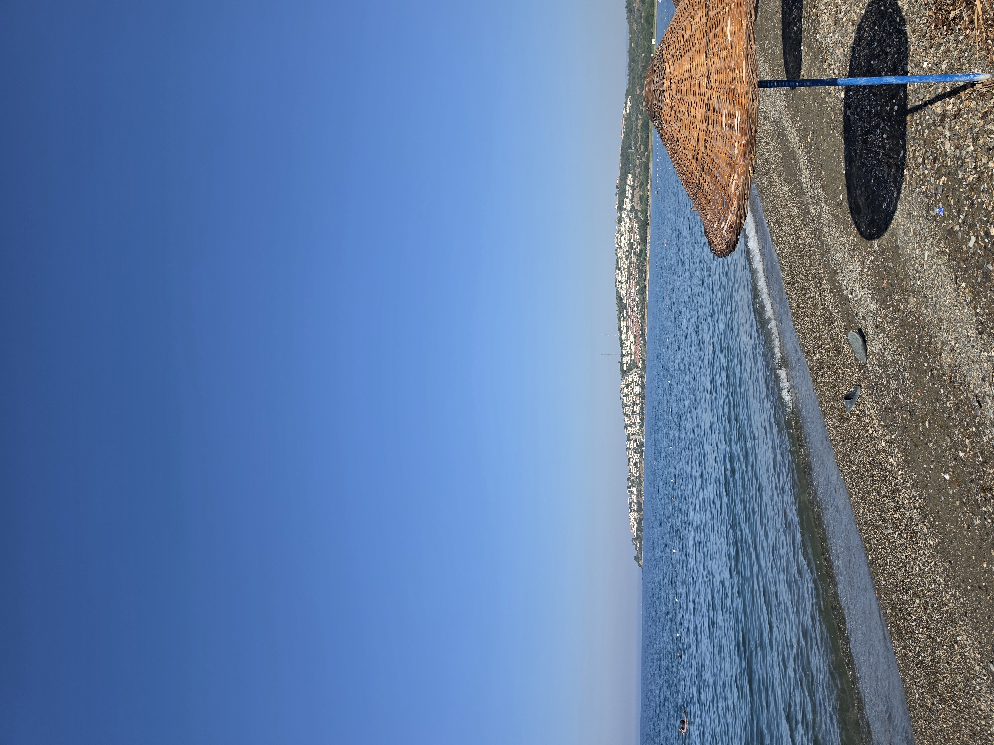 This is a photograph taken on a beach in Seferihisar, Turkey. The image captures a clear blue sky dominating the upper portion, with the calm, blue sea stretching across the middle ground. In the distance, along the coastline, a dense cluster of white buildings with red roofs cascades down a hillside, suggesting a developed coastal town.
In the foreground, the beach is composed of dark pebbles and sand, with a single woven beach umbrella, supported by a blue pole, casting a shadow. There is a dark-colored flip-flop near the water's edge. The sea is gently lapping at the shore, with a few small white buoys visible in the water. Further out, a person can be seen in the water, appearing to be swimming or relaxing on an inflatable. The overall scene depicts a tranquil day at the beach.