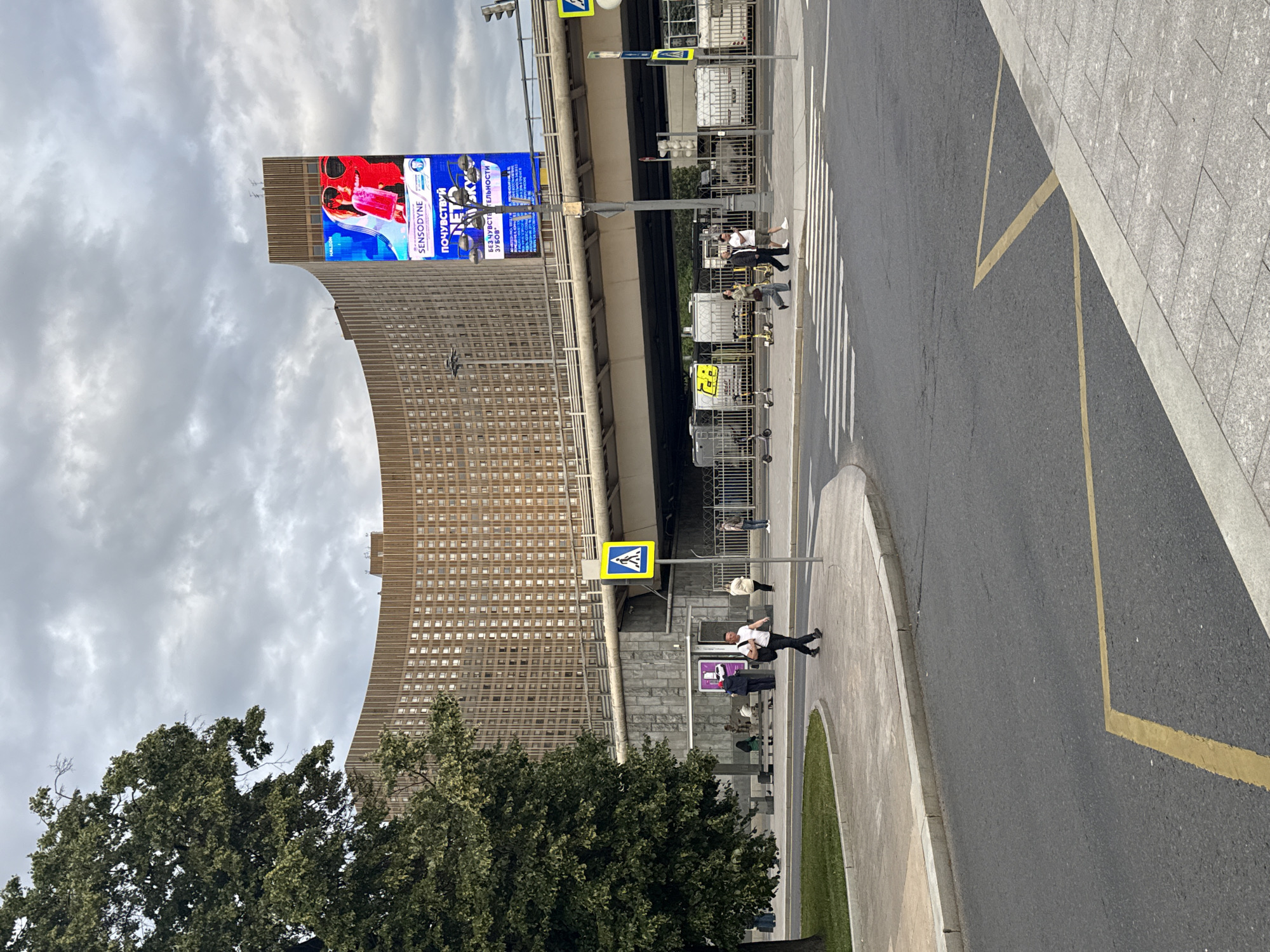 The image captures a wide street scene in Moscow, Russia, under a cloudy sky. A large, curved, brown-tiled building dominates the background, its many windows reflecting the overcast conditions. A digital billboard is mounted on the building's upper section, displaying an advertisement for Sensodyne toothpaste, with text in Russian that appears to say "ПОЧУВСТВУЙ НЕТ СКУ" and "БЕЗУПРЕЧНОЙ ТЕЛЬНОСТИ ЗУБОВ".
In the foreground, a street with pedestrian crossings is visible. Several people are scattered along the sidewalk and street. One person in a white shirt and dark pants walks across the street, while another in a white shirt and dark shorts is on the sidewalk. Further down the street, a few individuals are gathered, seemingly waiting or walking. Yellow pedestrian crossing signs are posted at intervals along the street.
The overall impression is a bustling urban environment, likely during daytime given the visibility and the nature of the activity. The cloudy weather contributes to a somewhat muted color palette, but the bright advertisement on the billboard adds a splash of color. The presence of the large building, street signs, and pedestrian crossings clearly indicates an urban setting.