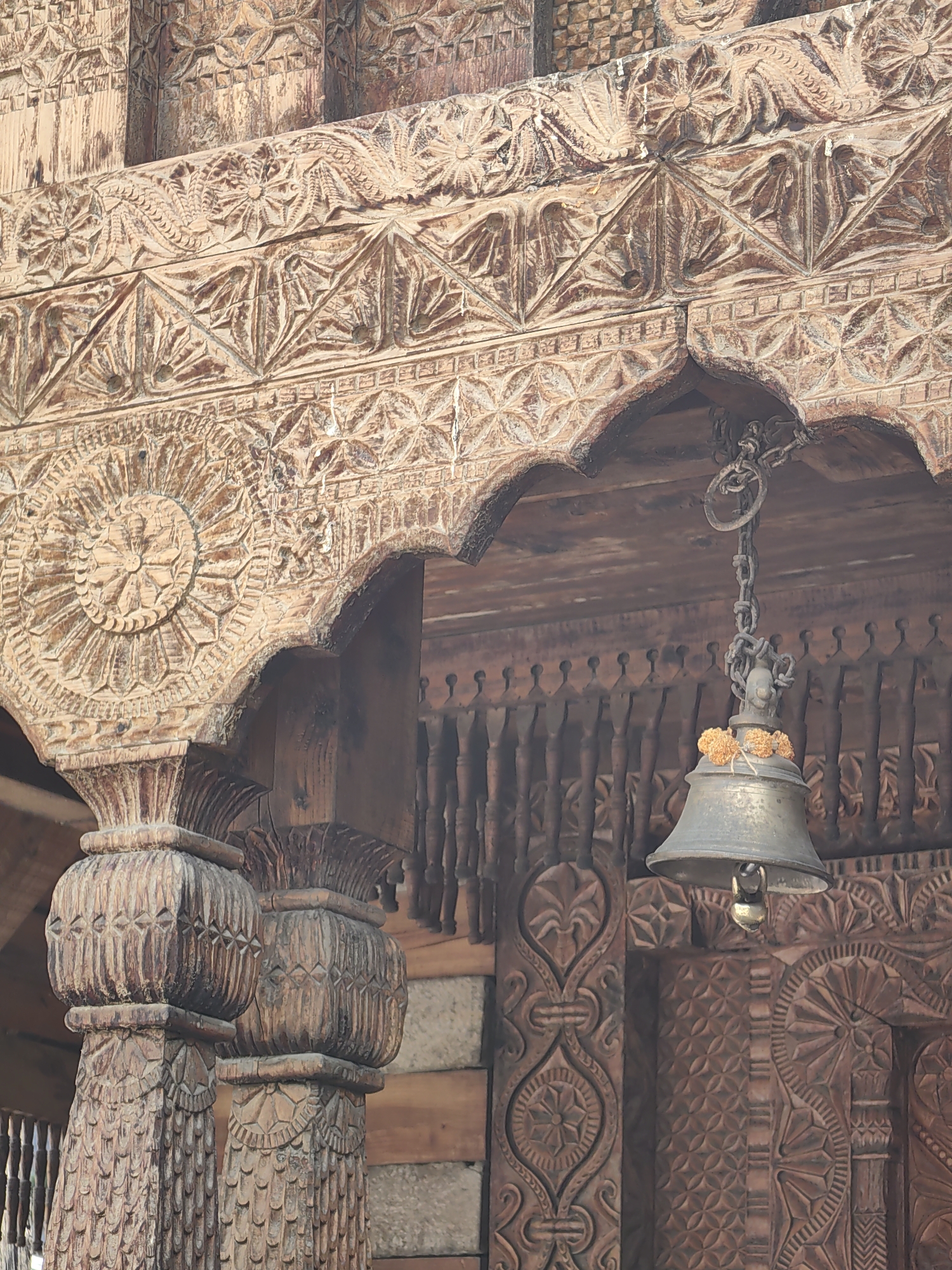The image displays intricate wooden carvings on the exterior of a building, likely a temple, in Vashisht, India. A bell, adorned with marigold flowers, hangs from a chain attached to the carved wooden structure. The detailed carvings feature geometric patterns, floral motifs, and architectural elements such as arches and columns. The overall impression is one of traditional craftsmanship and religious significance.