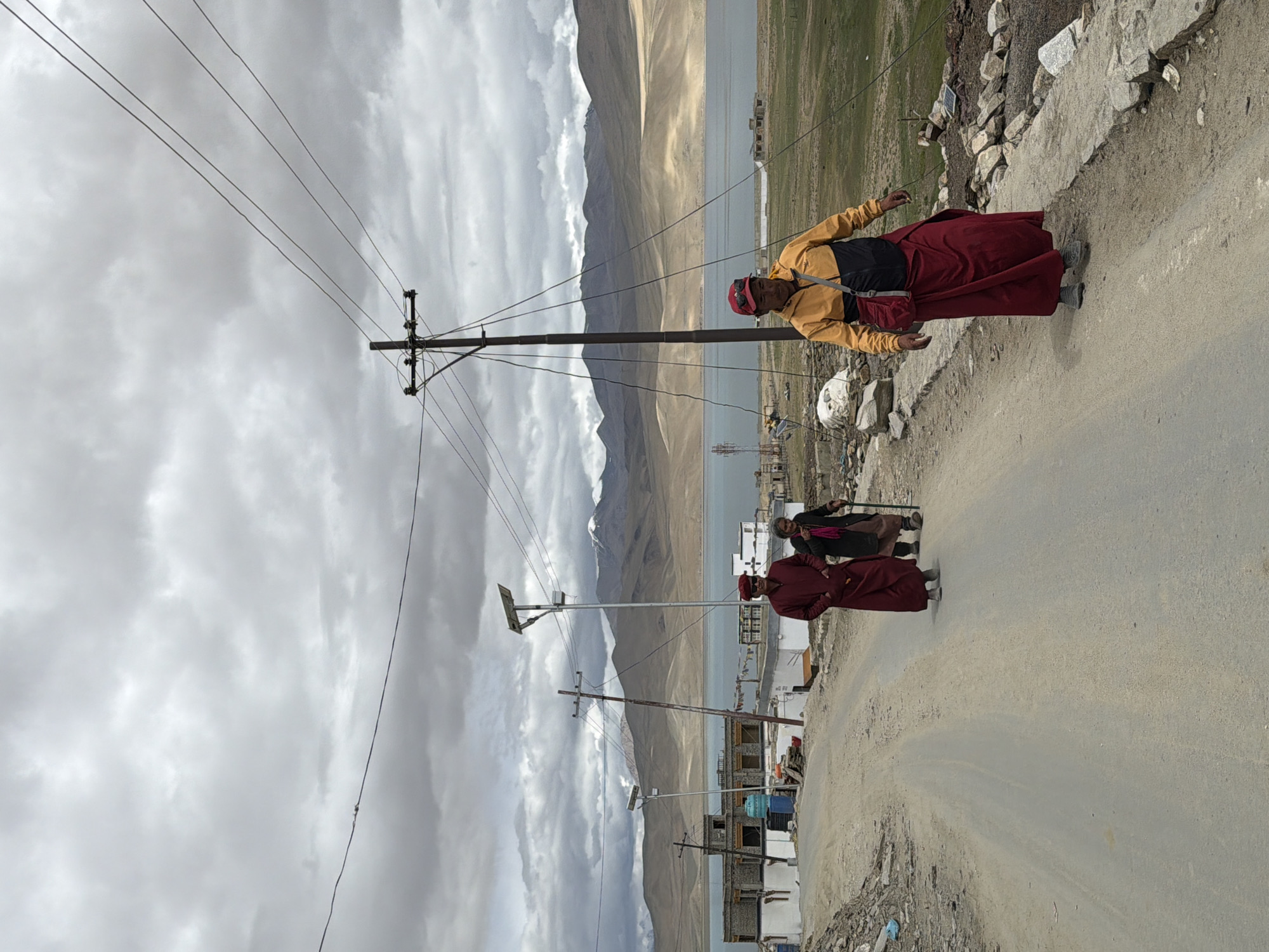 In the village of Karzok, India, three individuals are walking along a dusty road with a large body of water and mountains in the background. The sky is heavily clouded. The person in the foreground is wearing a yellow and black jacket over a long red robe, with a red bag slung over their shoulder. Behind them, two people in traditional maroon robes walk together. The person on the left is wearing a maroon robe, and the person on the right is wearing a darker robe and carrying a walking stick. Structures that appear to be buildings, some with white walls and others with brick, are visible on the left side of the road. There are also utility poles with wires stretching across the scene, and a solar panel mounted on a pole. The road itself is unpaved and appears to be made of gravel and dirt. The landscape beyond the road is a mix of green grassland and barren, mountainous terrain.