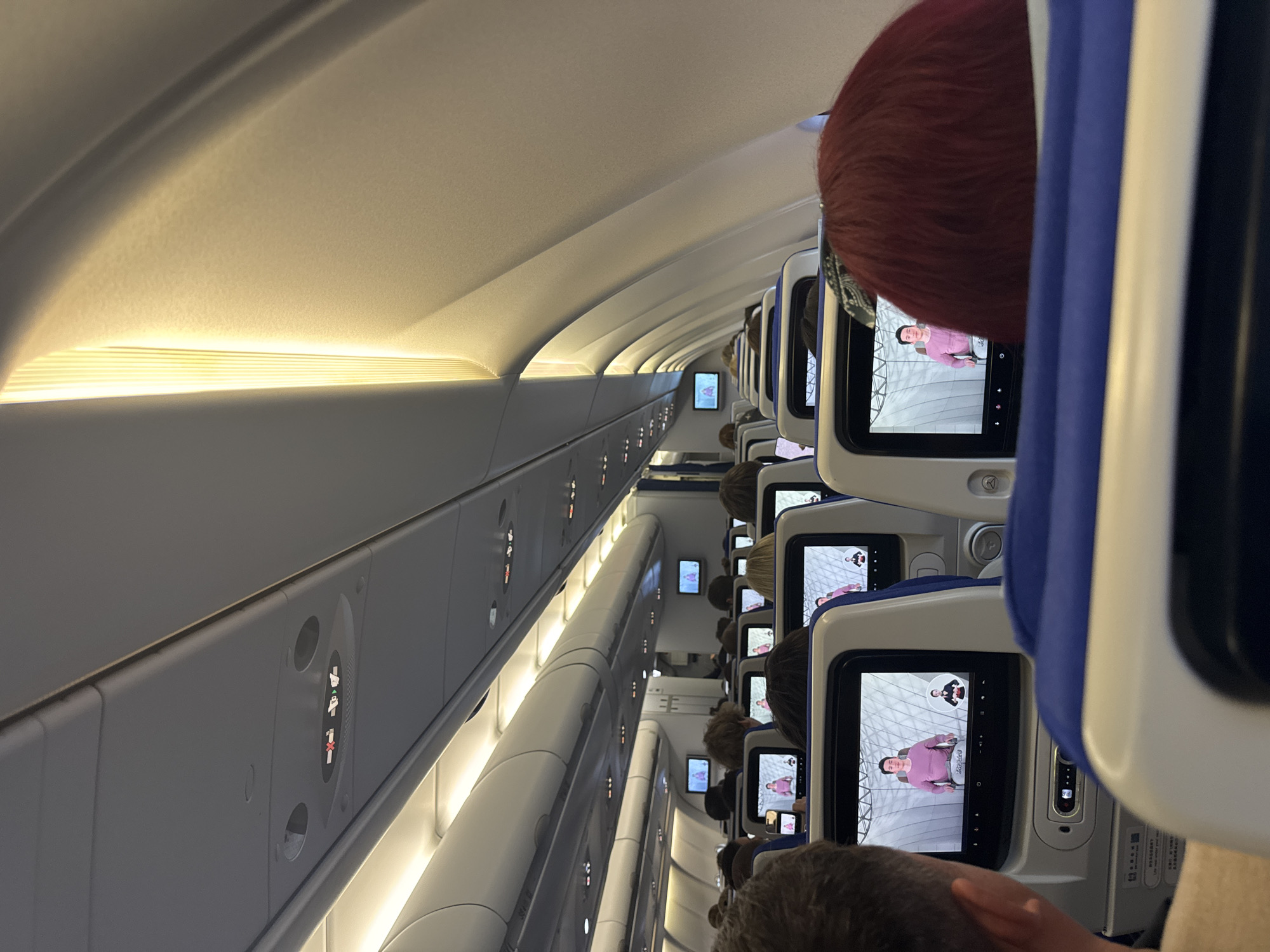 The image shows the interior of an airplane cabin, likely during flight. The cabin is filled with passengers seated in rows. Each seat has a personal entertainment screen displaying a video of a person in a pink shirt. Overhead, lighting illuminates the cabin. The scene appears to be in an airplane and no features indicate that the location is specifically Bol'shie Kabany, Russia.