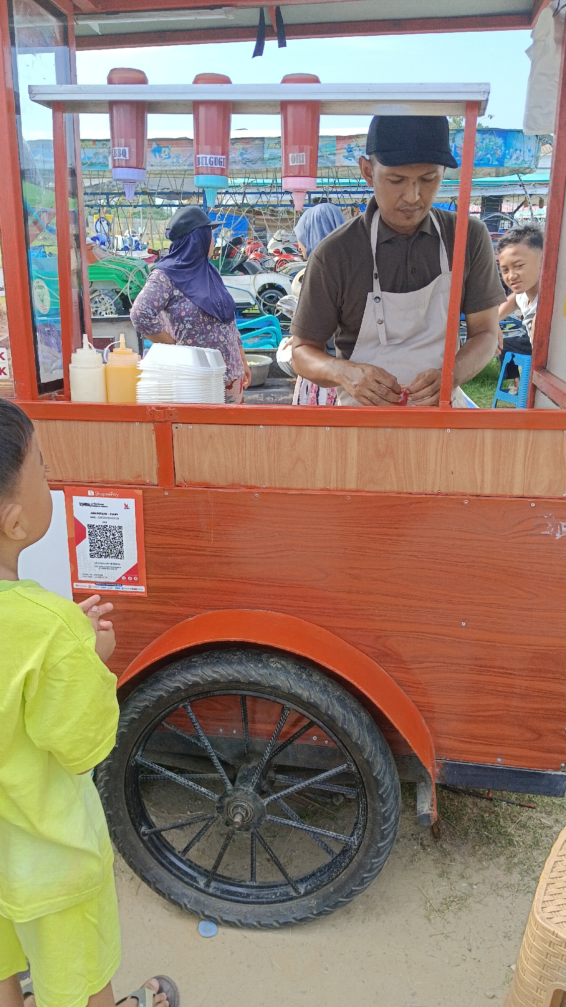 In Aek Kanopan, Indonesia, a food cart vendor operates under a bright, outdoor sky. The cart, constructed from reddish-brown wood with an orange trim, is equipped with a dispenser system holding three large containers labeled "BBQ," "BULGUGI," and "ORI," suggesting various sauce options. A man wearing a dark brown shirt, a white apron, and a black baseball cap is behind the counter, actively preparing food. To his left, a woman in a patterned purple dress and a dark hijab stands near a stack of white disposable containers and two squeeze bottles, one with a white liquid and another with a yellow liquid. On the far right, a young boy, partially visible, watches the scene. In the foreground, a young boy in a bright yellow t-shirt and shorts looks towards the cart. The cart features a QR code for payment, labeled "ShopeePay," prominently displayed on its side, below which is a wheel with a black rim and spokes. The ground appears to be sandy or dirt. In the background, an array of vehicles and structures suggest a lively outdoor environment.