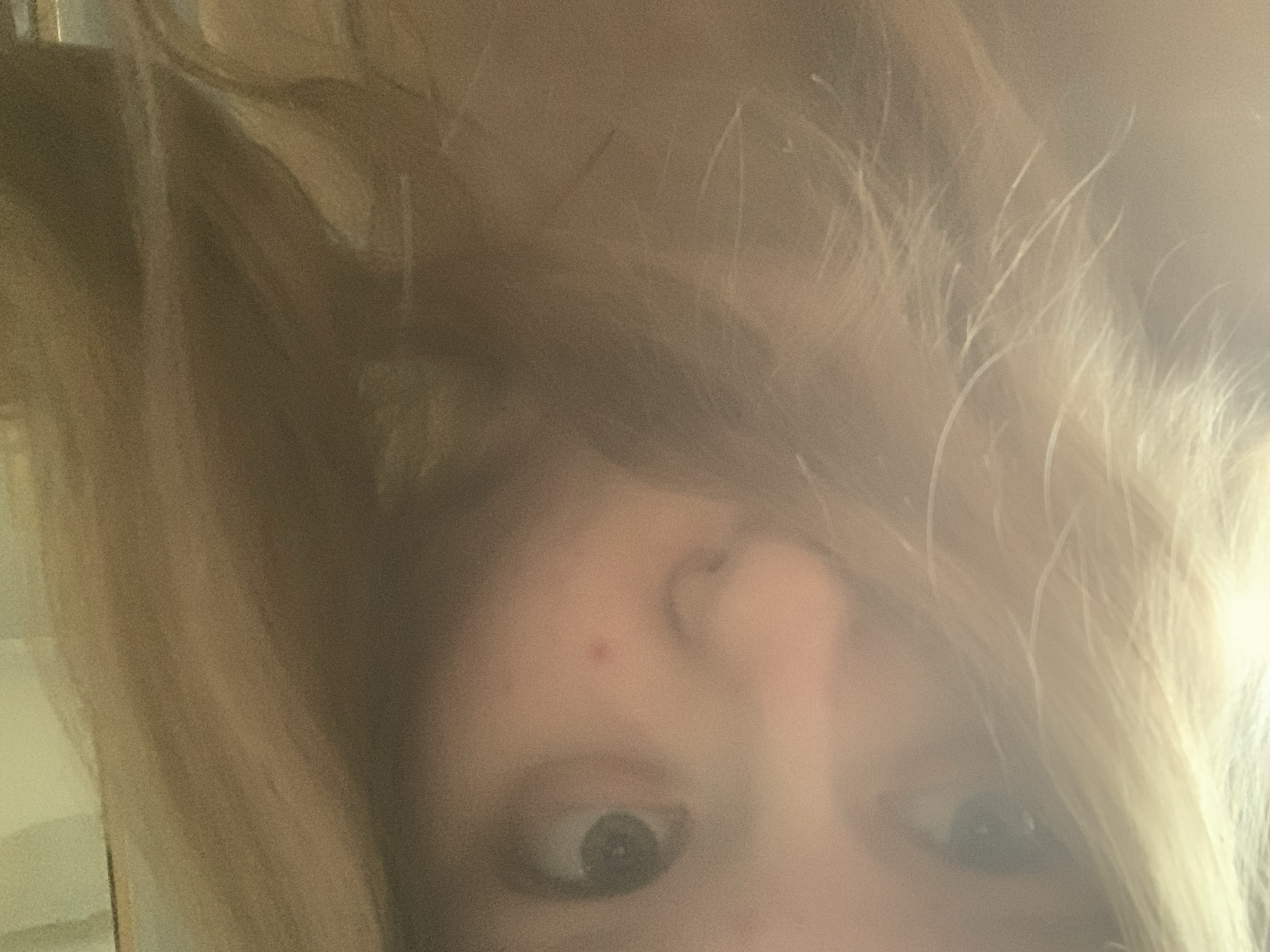 The media file is an inverted, close-up portrait of a person, likely taken as a selfie. The subject is a fair-skinned individual with light-colored, possibly blonde or light brown, hair that appears tousled and partially covers the face. The image is somewhat grainy and blurry, suggesting casual photography or challenging lighting conditions.
The most prominent features are the person's wide-open eyes, which are looking directly at the camera. The right eye (viewer's left) is more distinctly visible, showing a dark pupil and iris. The bridge of the nose is apparent, with a small red blemish or mole visible on the upper part of the nose or lower forehead. The left eye (viewer's right) is partially obscured by hair.
The image's orientation is upside down, presenting the hair as flowing upwards and the face positioned towards the bottom of the frame. The background is indistinct and out of focus, indicating an indoor setting without specific identifiable objects. The lighting is soft and diffused. No text is visible in the image. The wide-eyed expression conveys a sense of direct engagement or perhaps mild surprise. The location context for this personal capture is Saint Petersburg, Russia.