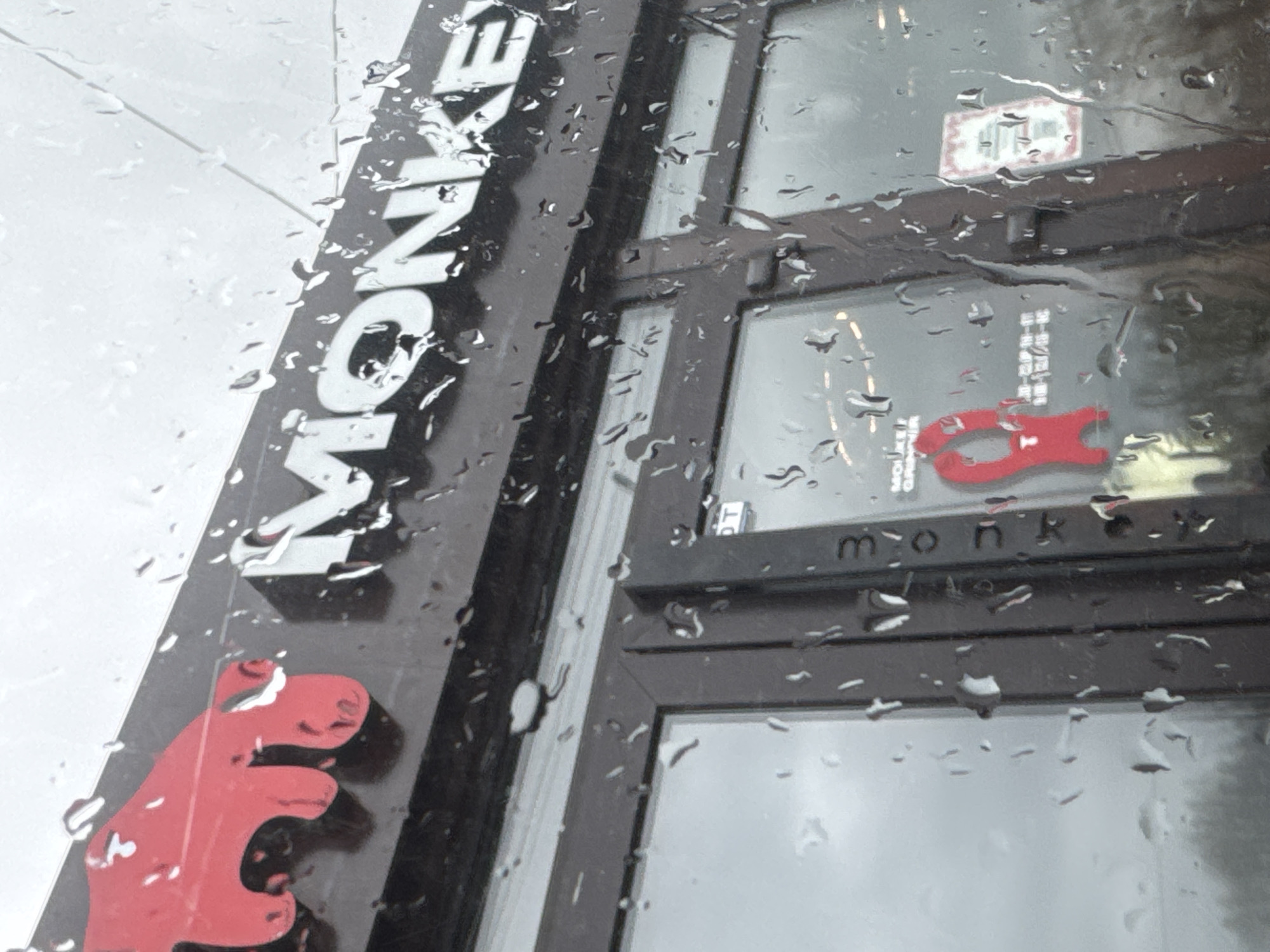 The photograph depicts the exterior of a "MONKEY" establishment, likely a business or shop, on a rainy day. The lettering is prominently displayed vertically on a dark facade, accompanied by a red animal logo resembling a monkey. The building has dark-framed windows, with glimpses of the interior visible through the raindrops on the glass. "monkey" is written in lowercase, stylized letters. The location is potentially in Perm, Russia, given the Russian characters observed inside.