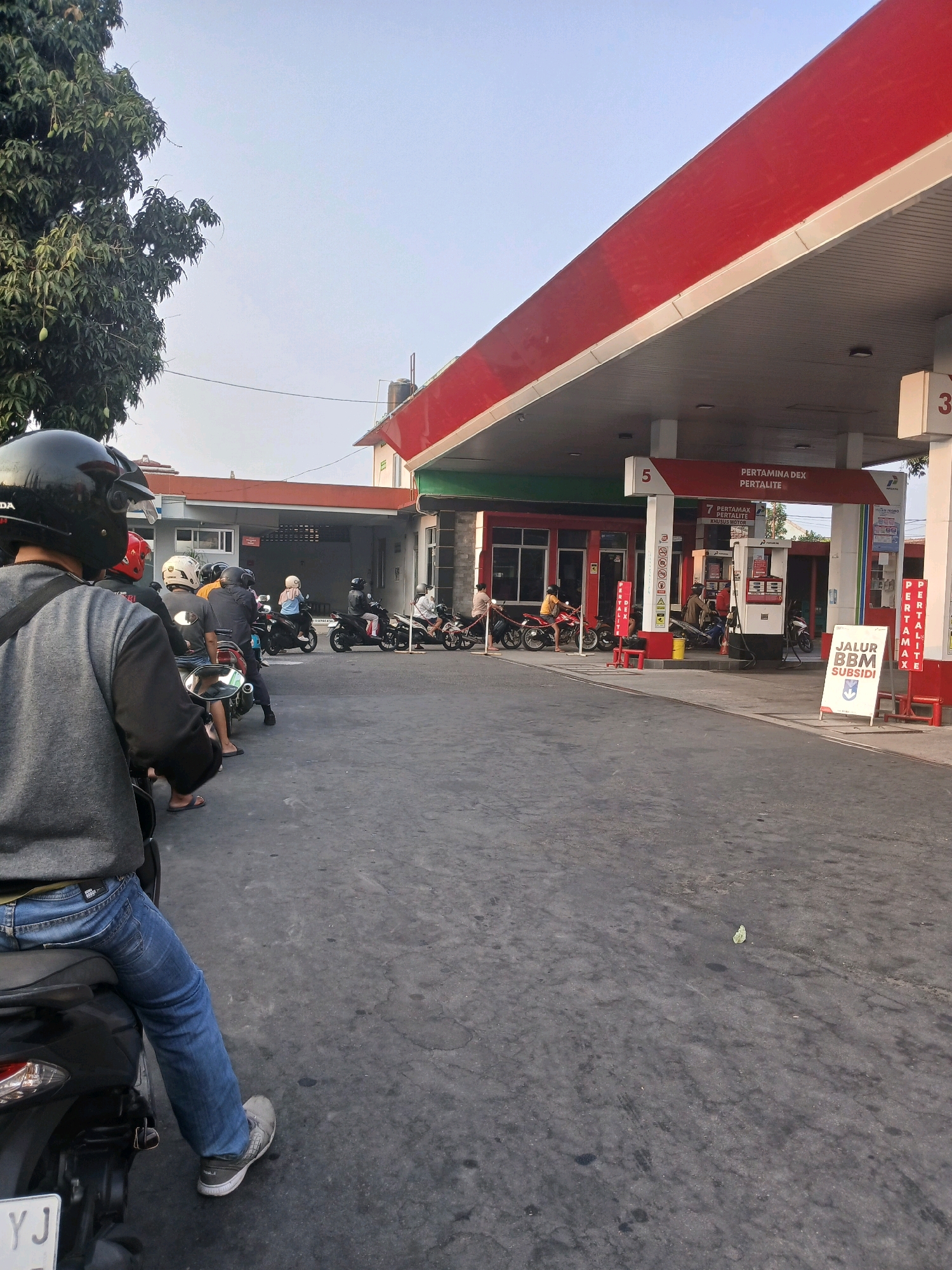 An outdoor medium shot captures a line of motorcycles waiting at a Pertamina gas station in Depok, Indonesia. The foreground is dominated by the back of a person wearing a gray jacket and blue jeans, seated on a black motorcycle, facing away from the camera. The motorcycle has a license plate that is partially visible. Behind this person, several other motorcycles and riders form a queue stretching towards the gas station's canopy. The gas station has a red and white color scheme, with prominent signage indicating fuel types like "Pertalite" and "Pertamax Dex". Several fuel pumps are visible under the canopy. In the background, a building with a red roof and white walls stands, and a large tree with green foliage is present on the left side of the image. The ground is paved with asphalt, showing some wear and tear. The sky is clear and bright, suggesting daytime. The scene appears to be a normal day at a busy gas station with customers queuing for fuel.