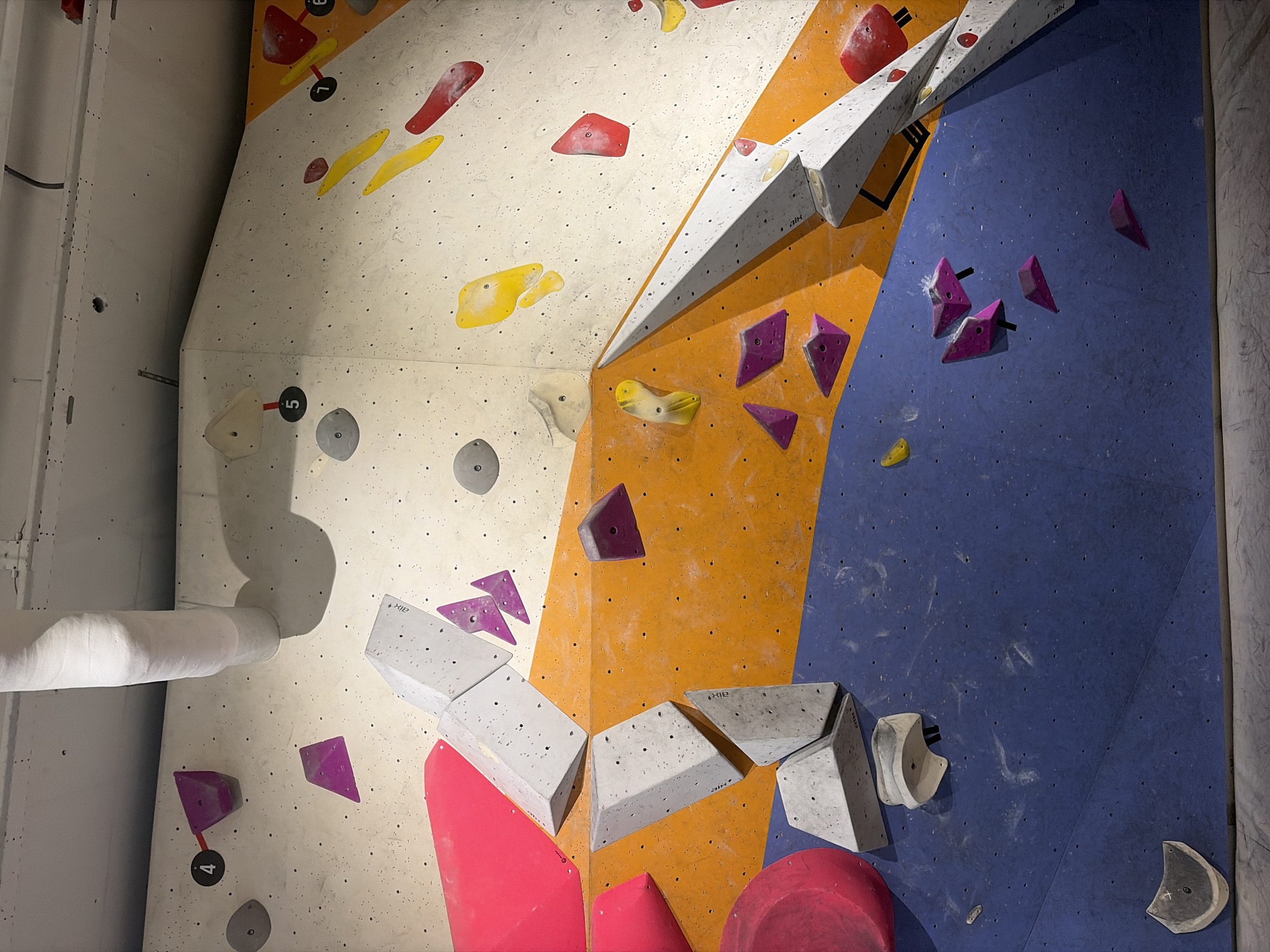 The image shows a section of an indoor climbing wall at an indoor climbing gym. The wall is constructed with various textured panels, primarily in white, orange, and blue. Numerous climbing holds in various shapes and colors, including pink, purple, yellow, red, and gray, are attached to the wall. Some of the holds are numbered, with visible numbers including 4, 5, 6, and 7. A large white pipe, possibly a ventilation shaft or structural element, is wrapped in white material and runs vertically along the upper left portion of the wall. The floor in front of the wall appears to be a padded matting typical of climbing gyms. There are no people visible in the image. The location is not discernible from the image itself, and therefore cannot be identified as being in The Hague, Netherlands.