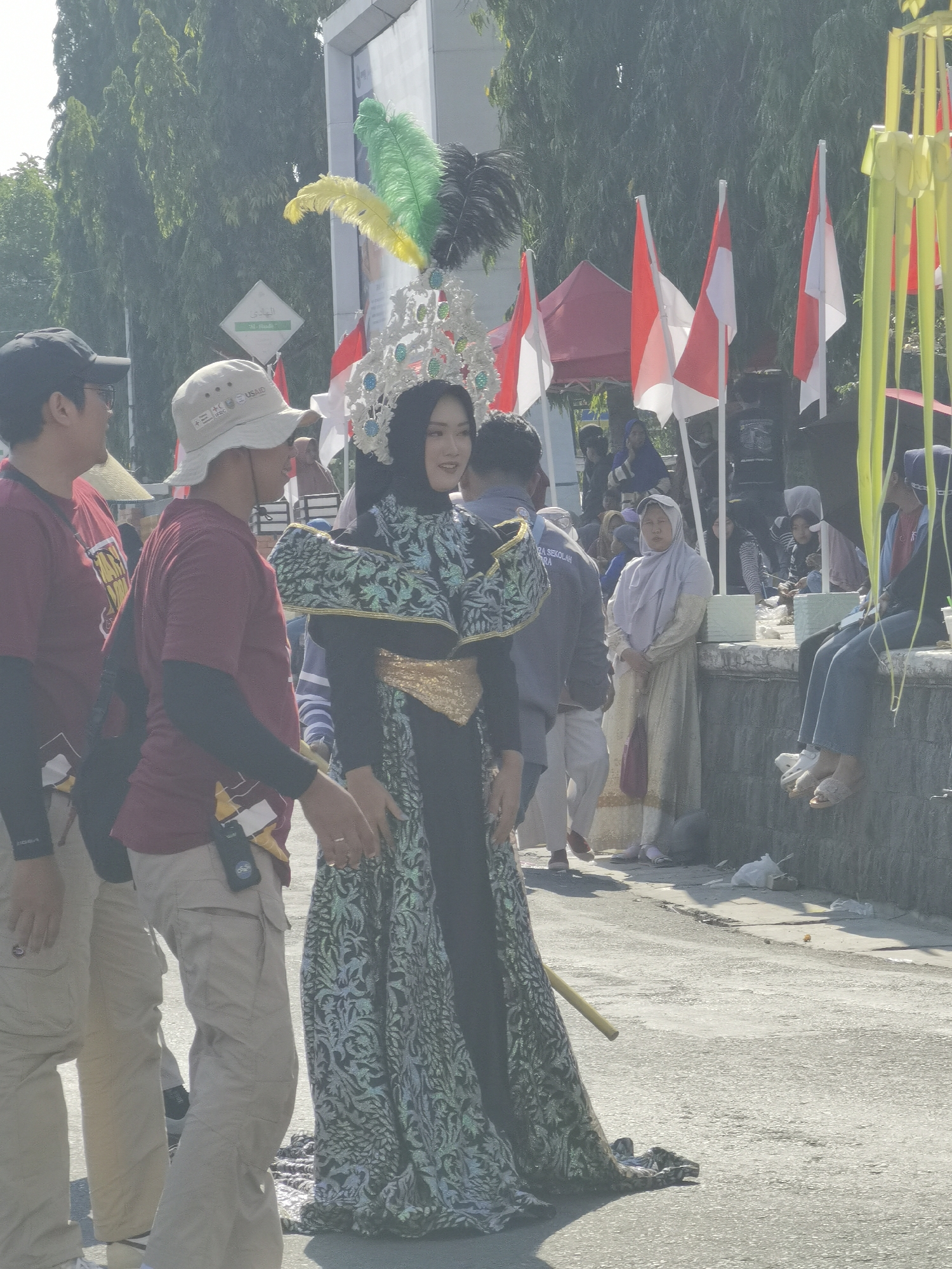 In the city of Blora, Indonesia, on a sunlit day, a group of people are gathered outdoors. In the foreground, a woman adorned in a black and green ornate dress and a jeweled headdress with feathers, stands prominently. She appears to be part of a procession or performance. To her left, two men in casual attire, one wearing a maroon shirt and a bucket hat, are visible. The man in the bucket hat has a USAID logo on it. Behind them, several Indonesian flags with red and white stripes flutter in the breeze. In the background, more people are milling about, some seated on a low wall, and a red tent is partially visible. The environment suggests a public event or festival taking place on a street or walkway.