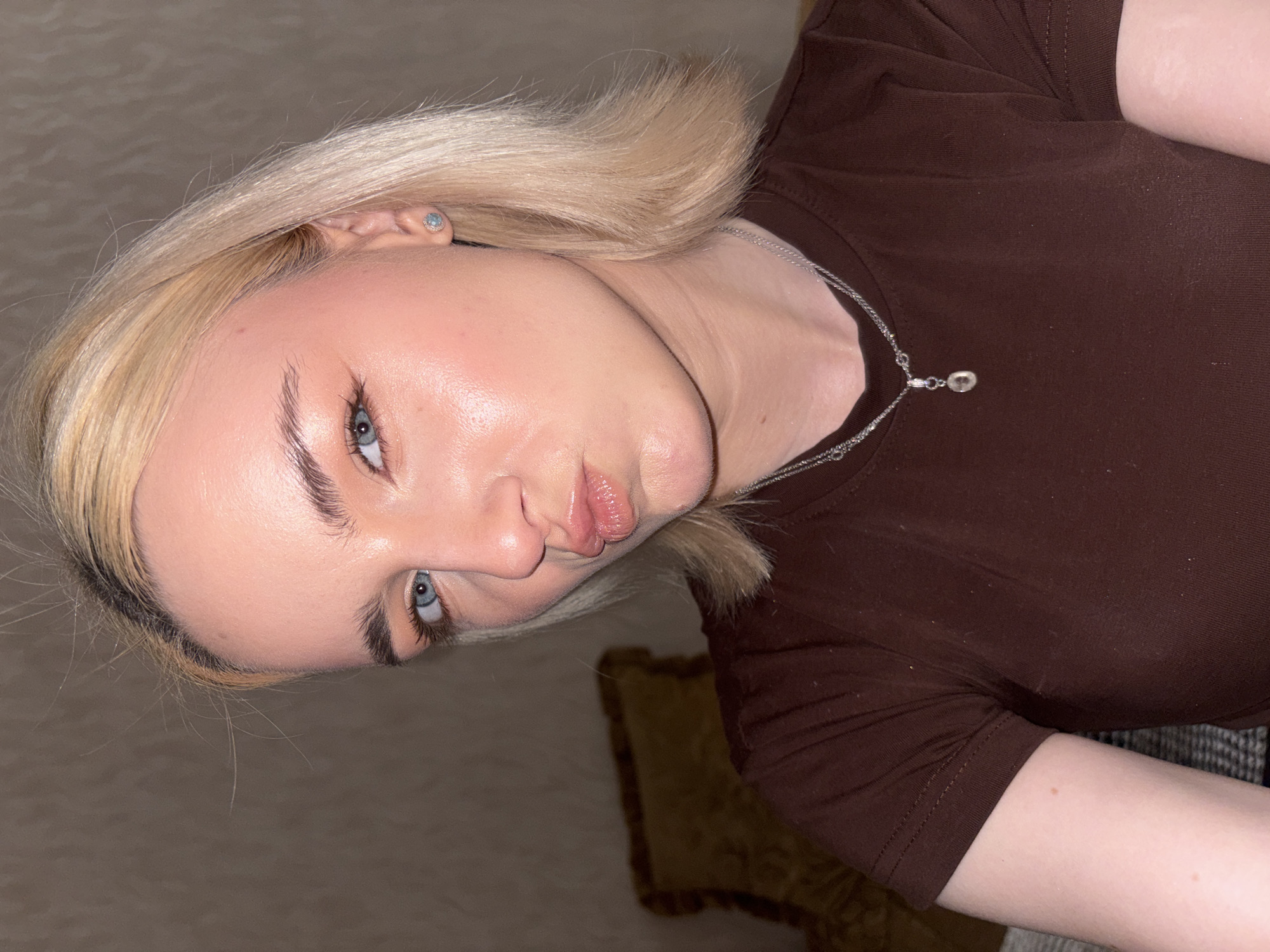 A young woman with blonde bobbed hair and blue eyes is pictured in a close-up portrait. She is wearing a brown t-shirt and a silver necklace with a pendant. Her lips are puckered, and she has on light makeup, including blush and eyeliner. A diamond stud earring is visible on her right ear. The background is a plain, textured wall. There is no indication of the location being in Sinyavino, Russia.