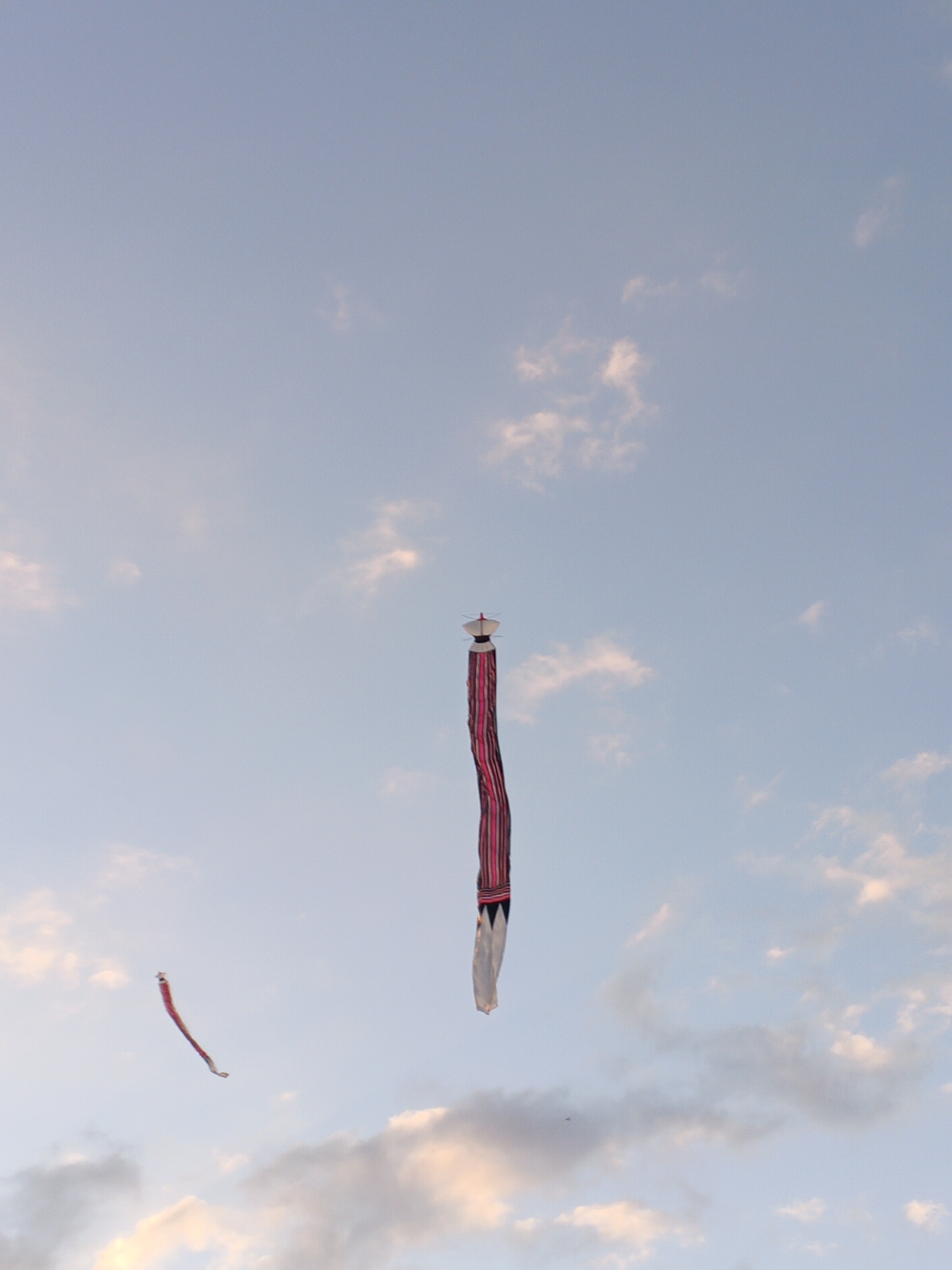 In the city of Sukawati, Indonesia, two kites fly against a pale blue sky dotted with wispy white clouds. The larger kite, positioned centrally and vertically, features a white top and a long, striped tail of pink and dark red. To the left and slightly lower, a smaller kite, also appearing to have a reddish hue, is suspended in the air, angled diagonally. The image captures a tranquil outdoor scene, likely during daylight hours, with the focus on the kites in flight against the expansive sky.