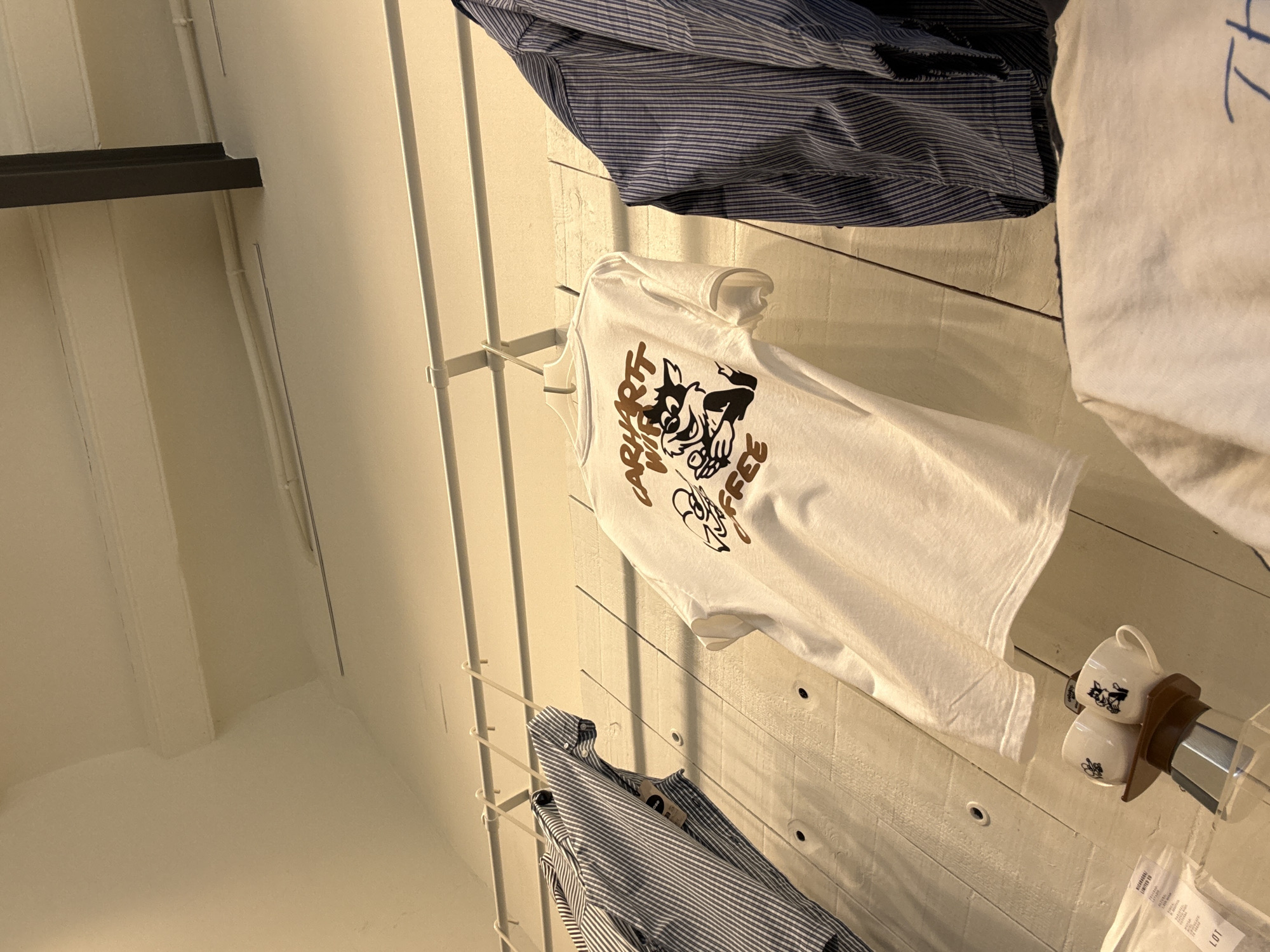 The image shows an interior scene, possibly a retail space or a workshop, with clothing items displayed on hangers against a white wooden wall. In the foreground, a white t-shirt with a graphic design featuring a cat character holding a coffee cup and the text "CARHARTT WIP COFFEE" is prominently displayed. To the left, a striped shirt is hung, and to the right, another striped garment is partially visible. Below the t-shirt, two small white coffee cups with illustrated designs, possibly matching the t-shirt graphic, sit on a brown wooden stand next to a metallic coffee maker. The background reveals a white ceiling and a structural beam. The overall impression is of a curated display of merchandise. The location is identified as Barcelona, Spain, suggesting this is likely a shop or a design studio. There are no people visible in the image.