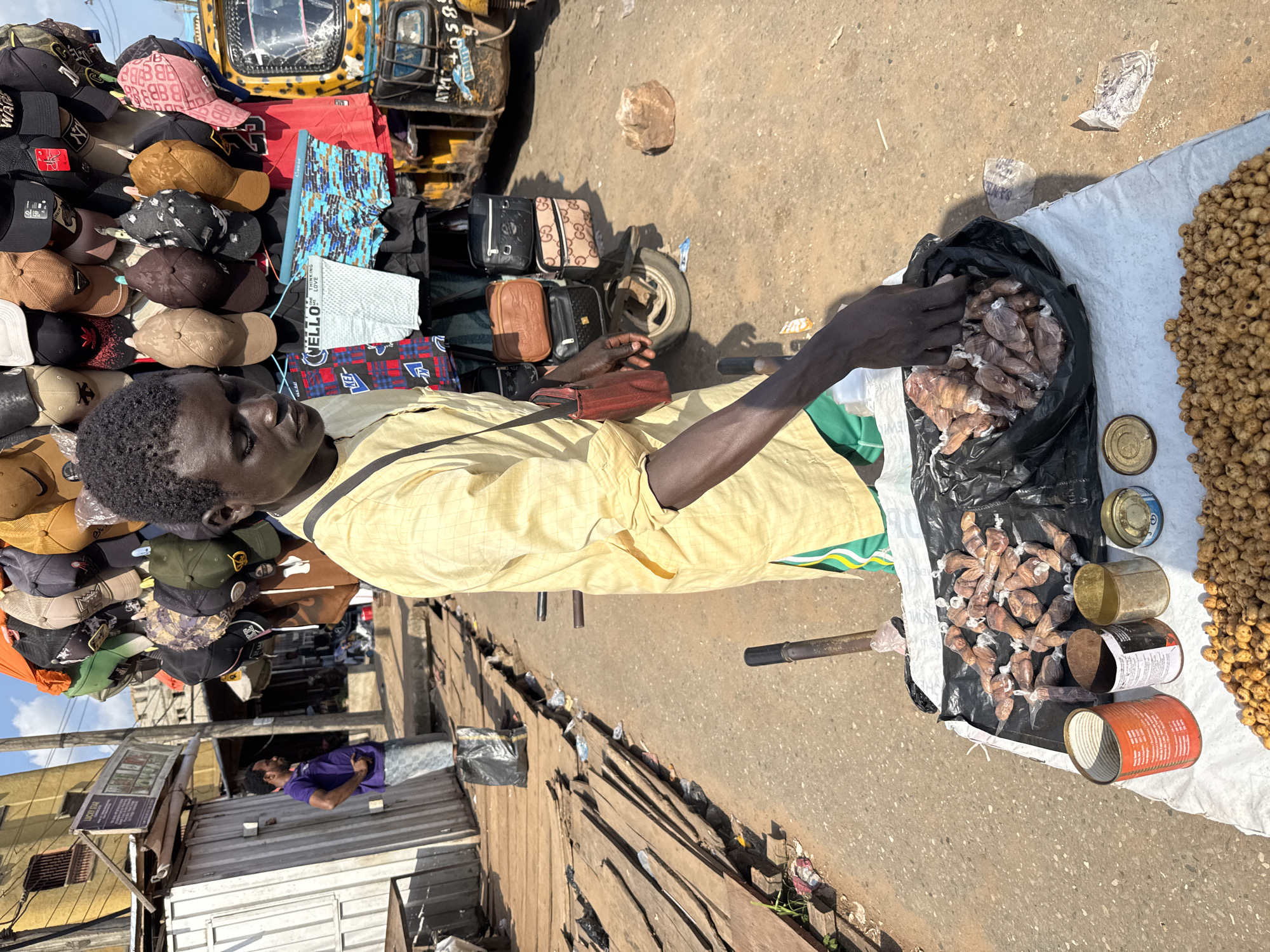 An adult male in Nkpor, Nigeria, wearing a light yellow collared shirt with black trim is selling goods on the street. He is standing at a white cloth covered surface which has a bag of small wrapped items, several open metal cans, and a mound of tan, globular items. Behind the seller is a collection of hats, bags, and clothing attached to a yellow vehicle with a broken windshield. The street surface is adjacent to a weathered wooden structure. In the background a man in a purple shirt stands by a small building.