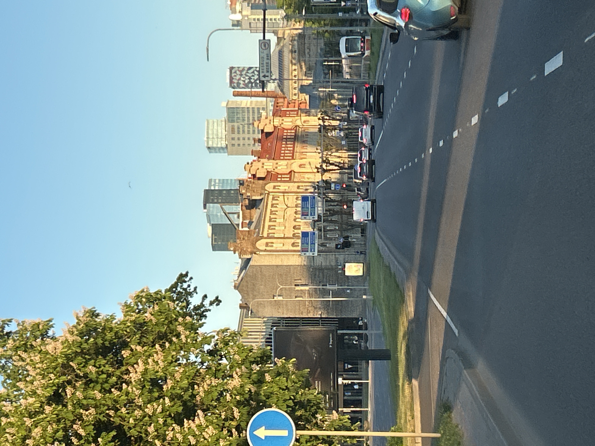 The image depicts an urban street scene in Tallinn, Estonia, specifically on or approaching Väike-Pärnu maantee, looking towards the Kesklinn (city center) and Ülemiste district. The lighting suggests late afternoon or early evening, characterized by long shadows stretching across the road, originating from the west. The sky is clear and bright blue.
In the immediate foreground on the left, a large, leafy tree with numerous clusters of light-colored blossoms, likely a horse chestnut, partially obscures the view. Below the tree, a blue circular traffic sign with a white upward arrow indicates a "straight ahead" mandatory direction. Adjacent to the tree, a black billboard is visible but mostly obscured by shadow and the tree itself.
The asphalt road, comprising multiple lanes, stretches into the distance, marked by dashed white lines. Several vehicles are present on the road, all moving away from the viewer. These include a white panel van, a black SUV, and at least three white sedans or hatchbacks. On the far right, partially visible, is the rear section of a light blue/teal car, possibly a compact SUV, with its taillights reflecting the sunlight.
Along the right side of the road, a modern white and red tram is visible on its tracks, appearing to be stationary or moving slowly. Further down the road, traffic lights show red. A directional street sign above the road indicates "KESKLINN, ÜLEMISTE" (City Center, Ülemiste) and "VÄIKE-PÄRNU MNT".
The background features a blend of architectural styles. On the left, a grey concrete building stands next to older structures. Further into the distance, a row of multi-story buildings with distinct reddish brick facades and roofs are visible. Beyond these, the modern skyline of Tallinn emerges, with several glass-clad high-rise buildings and one distinctive tall building with a patterned facade. A faint, dark, bird-like object is visible in the upper sky to the left. The overall scene captures a vibrant urban environment during golden hour.