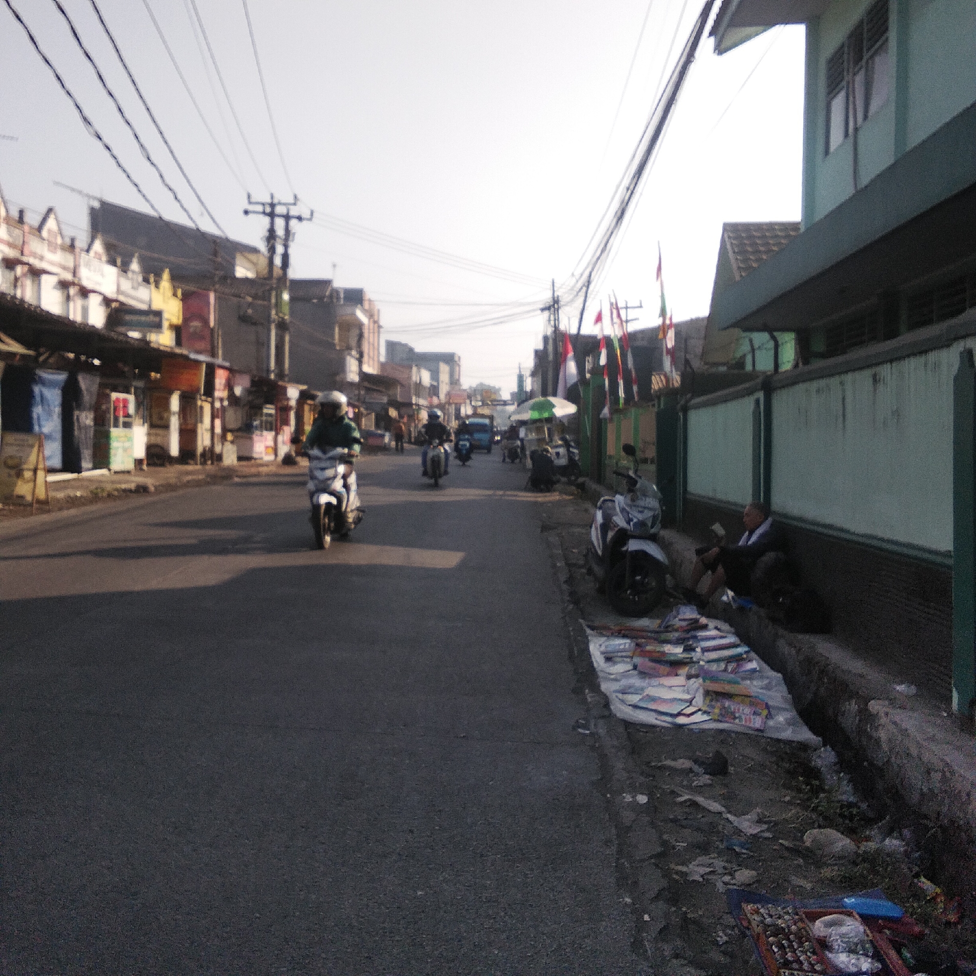 In Cisaat, Indonesia, on a street lined with buildings and small shops, a man is seated on the ground beside a pile of items laid out on a tarp. He is wearing dark clothing and appears to be looking at something in his hands. Next to him, a motorcycle is parked. The street is paved and appears to be a commercial area, with several other motorcycles visible in the background, some with riders. Flags are displayed on poles along the side of the street. The buildings on the left appear to be shops or residences with awnings and signage. The sky is overcast and bright.