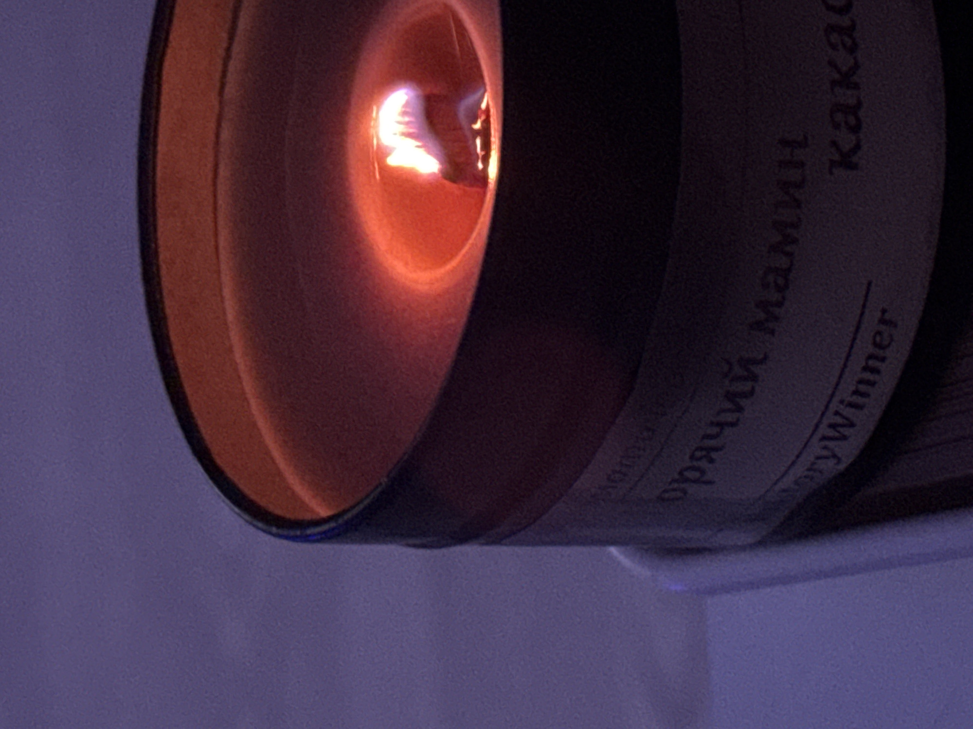 The image shows a close-up of a burning candle. The flame is visible within the candle's well, casting a warm, orange glow. The side of the candle jar is visible, with text in Cyrillic script that includes the words "мамин" and "какао," suggesting a cocoa-scented candle. Part of the English text "oryWinner" is also visible. The background is a blurry purple color, likely a wall or surface the candle is placed near. There is no information in the image that indicates a location in Ново-Никольское, Russia.