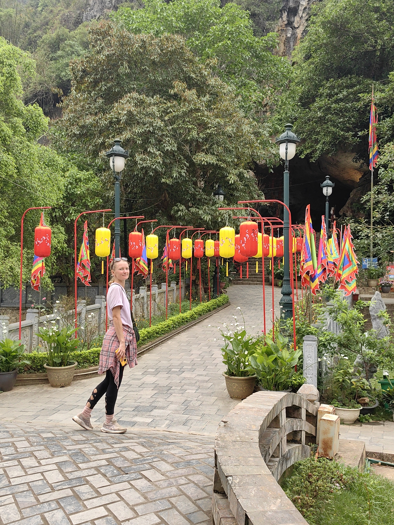A woman stands on a brick pathway surrounded by lush greenery and decorations in Lạng Sơn, Vietnam. She is fair-skinned with blonde hair, wearing a light pink t-shirt, black leggings with cut-out designs, and pink sneakers. A plaid shirt is tied around her waist. She is facing the camera.
The pathway is made of gray bricks and is lined with red metal frames from which hang red and yellow lanterns. The area is also adorned with colorful flags.
The background features a variety of trees and plants, with a rocky hillside visible above. Lampposts line the walkway. A stone bridge is visible in the foreground.