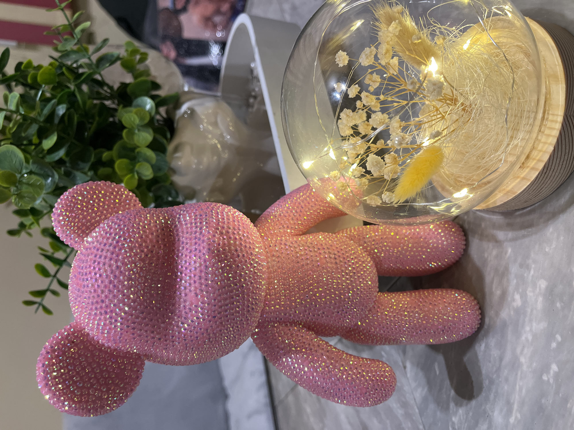 A close-up shot displays a pink, glitter-covered teddy bear lying on a light-colored surface. Beside it, a plant with small green leaves is visible, along with a glass dome containing dried white flowers and small yellow accents, illuminated by fairy lights. A white cylindrical object is also in the background. The scene does not provide enough information to determine the location as Veliko Tarnovo, Bulgaria.
