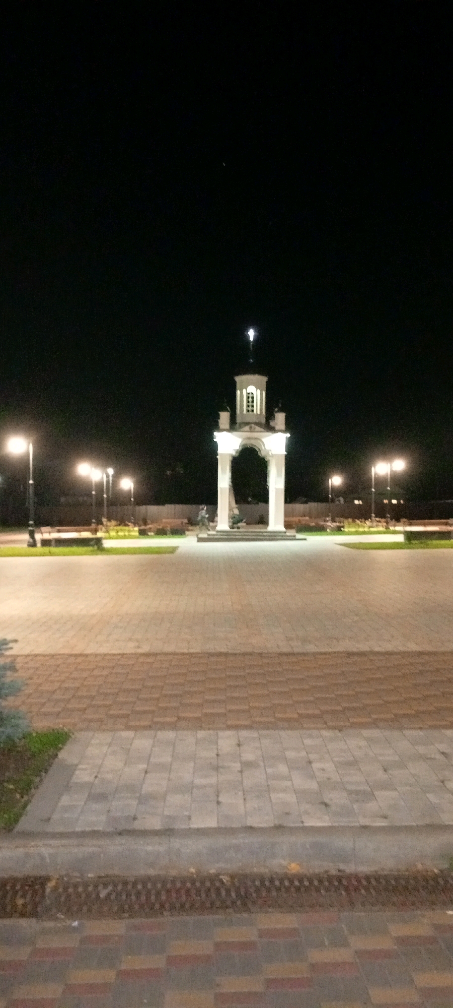 The image depicts a nighttime scene in a city park, likely in Kirsanov, Russia, centered around a white, arch-shaped monument topped with a cross. The monument is illuminated, casting a soft glow on the surrounding paved area. Several streetlights line a pathway to the left of the monument, also illuminated, and contribute to the overall lighting of the scene. In the background, beyond the paved area and illuminated monument, is a dark expanse, likely representing the night sky, with some distant lights suggesting the presence of buildings or further urban development. The ground in the foreground is paved with interlocking bricks in a patterned arrangement of grey and reddish-brown tones. A low curb separates the foreground paving from a strip of grass, which in turn borders the main paved area. A few small evergreen plants are visible in the lower left corner. The scene is devoid of any people, animals, or overt activity, presenting a serene and still urban landscape.