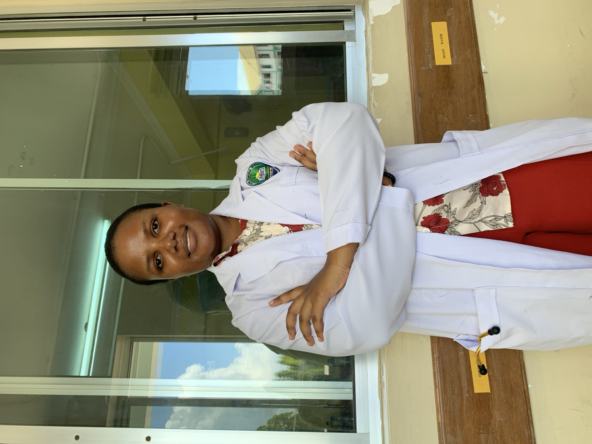 A woman wearing a white lab coat is standing indoors. She has her arms crossed and is smiling. She is wearing a red top with a floral pattern underneath her lab coat. She is standing in front of a window with a reflection of a building and sky. There is a sign labeled "SOAPY WATER" on the wall to her right. Based on the logo on the lab coat, the location is likely a university or educational institution in Dar es-Salaam, Tanzania.