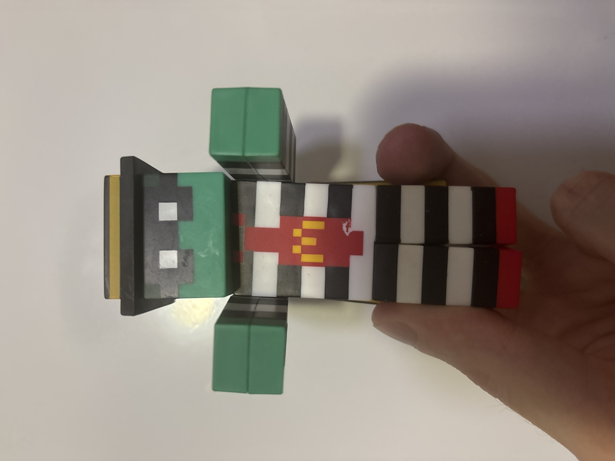 A person is holding a stylized toy resembling a character from the game Minecraft. The toy is green with a black hat that has a yellow band. It wears a black and white striped shirt with a red chest plate featuring a yellow McDonald's logo. The character has green block-like arms and red block-like feet. The background is a plain white surface.